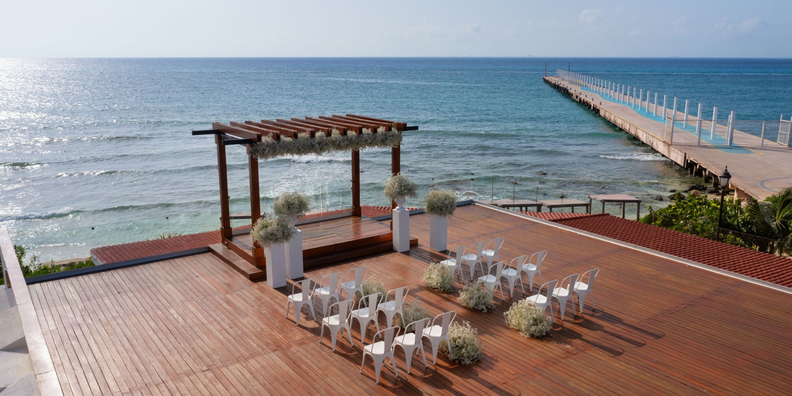 The Hilton Playa del Carmen provides a wooden deck overlooking the ocean, elegantly arranged for an outdoor event featuring white chairs and floral decorations beneath a pergola.