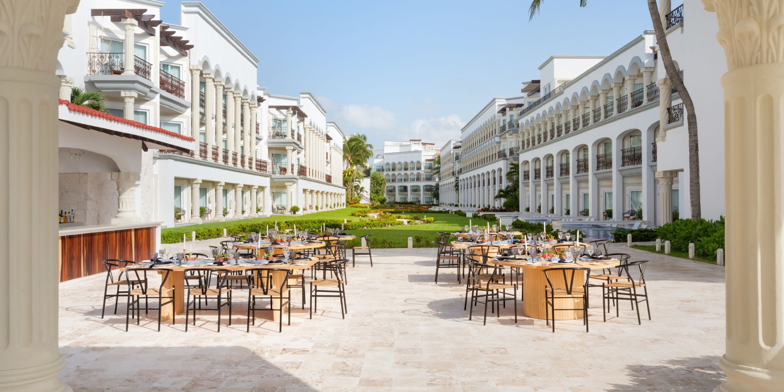 The Hilton Playa del Carmen features an inviting outdoor dining area with multiple tables elegantly set for a meal, nestled in a courtyard surrounded by light-colored buildings and lush greenery under a clear blue sky.