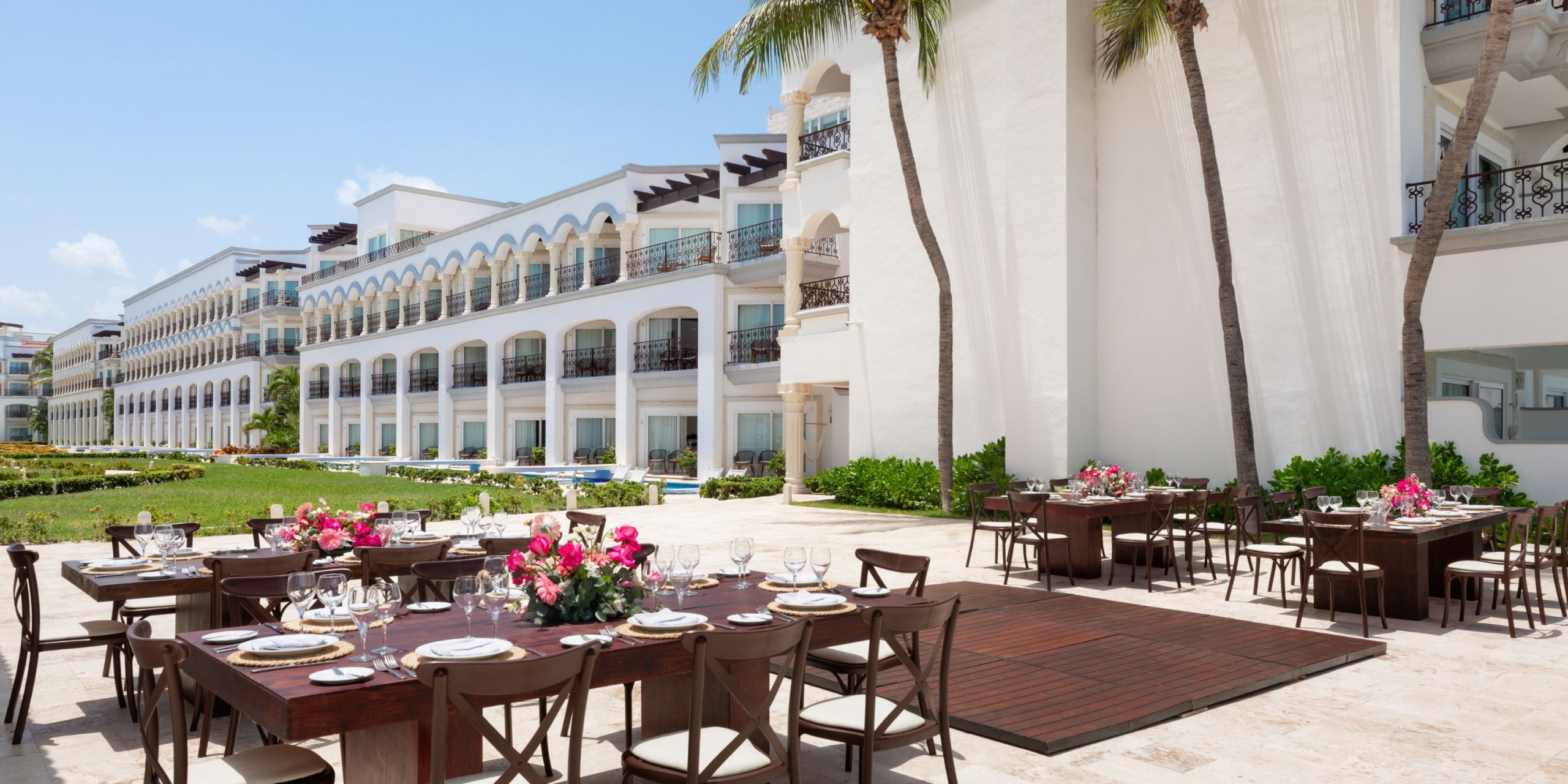 Enjoy an outdoor dining experience at the Hilton Playa del Carmen, featuring wooden tables elegantly arranged for an event with floral centerpieces. The white resort buildings with balconies provide a stunning backdrop, surrounded by lush palm trees and greenery.