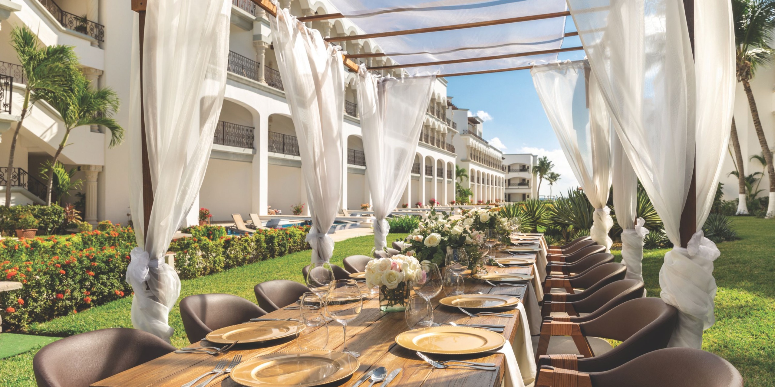 Under the pergola at Hilton Playa del Carmen, an outdoor dining setup showcases a long wooden table adorned with floral centerpieces, accompanied by brown chairs and framed by white curtains, all set against the lush backdrop of the hotel’s balconies and greenery.