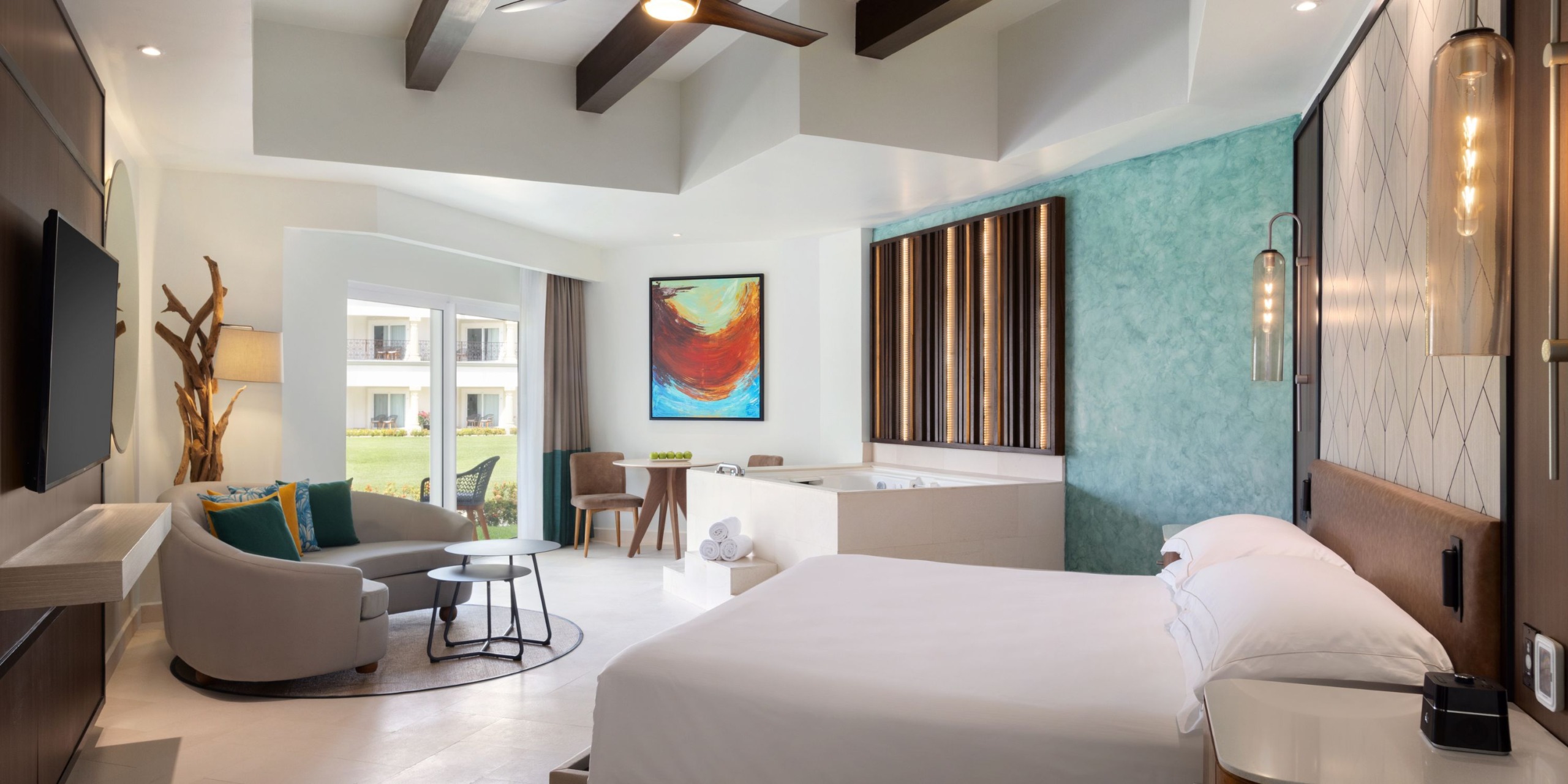 Experience a modern room at Hilton Playa del Carmen, featuring a king-sized bed, abstract artwork, a cozy seating area, a flat-screen TV, and views through a large window. The decor elegantly blends teal and neutral tones.
