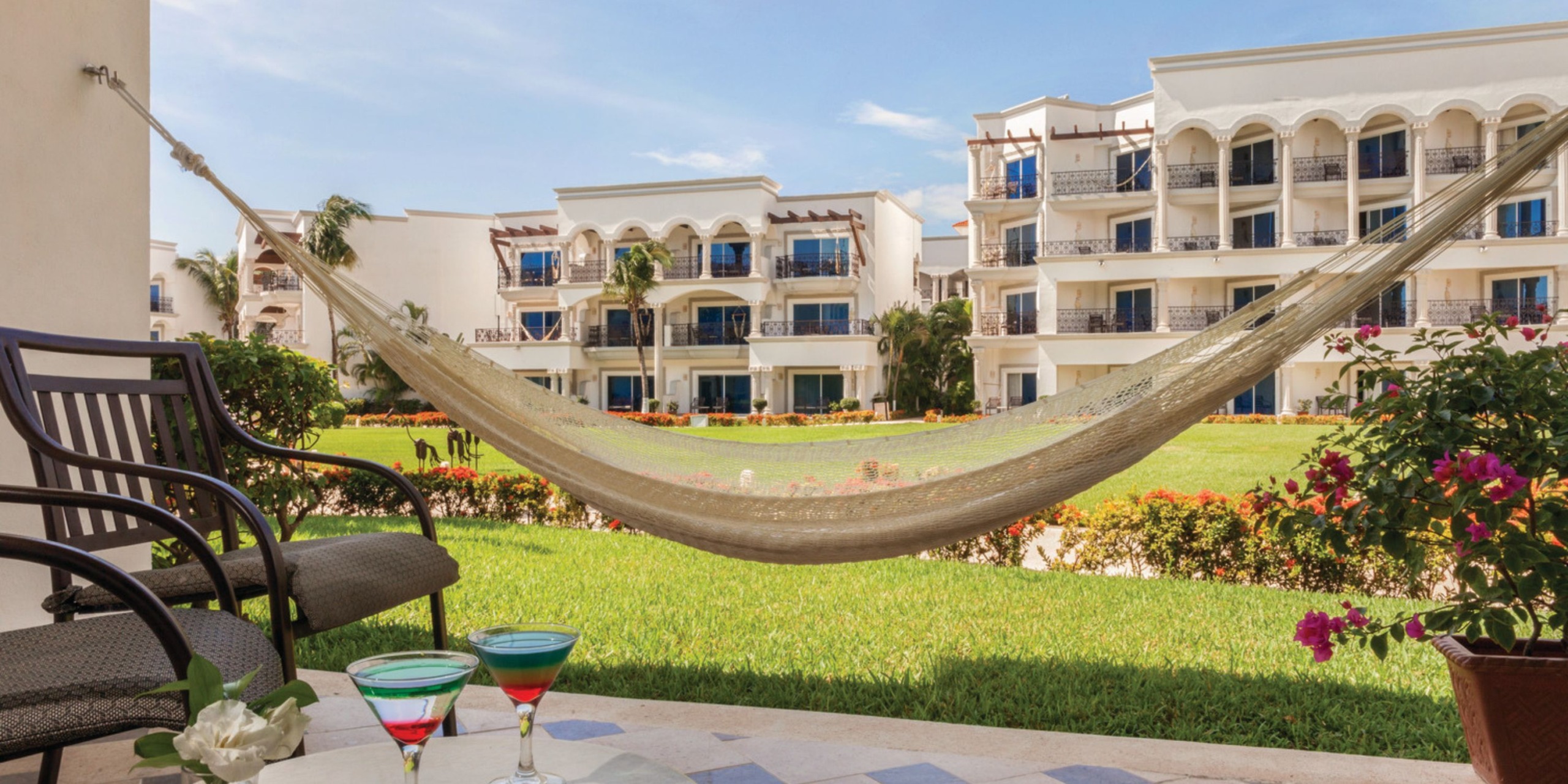 Enjoy a view from the Hilton Playa del Carmen patio, complete with a hammock, two chairs, and two cocktails, all overlooking a lush grassy area with resort buildings in the background.