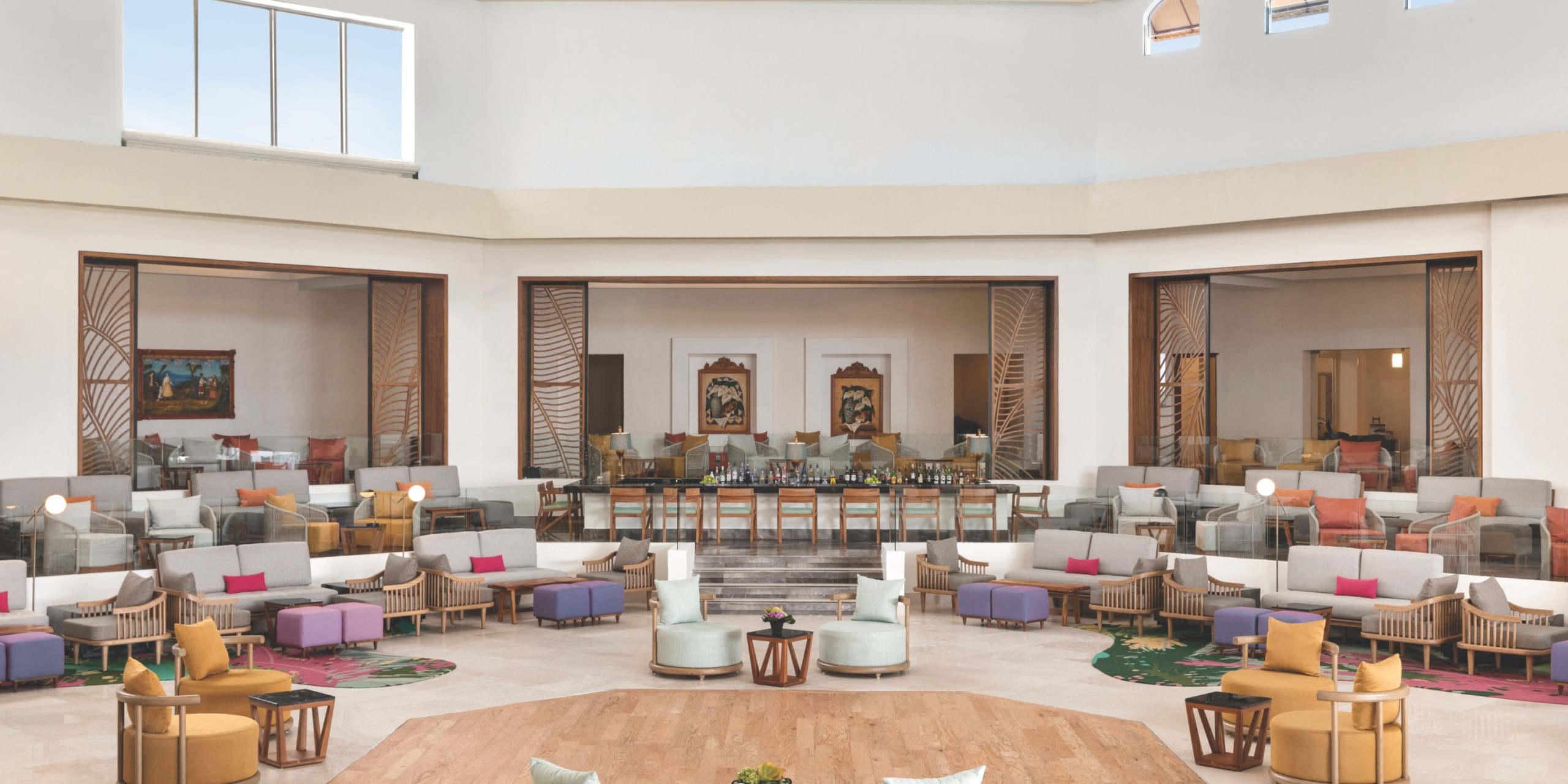 The Hilton Playa del Carmen features a spacious lobby area adorned with modern seating, vibrant cushions, and large windows. A central bar area, visible from various seating arrangements, enhances the atmosphere, while artwork elegantly decorates the walls.