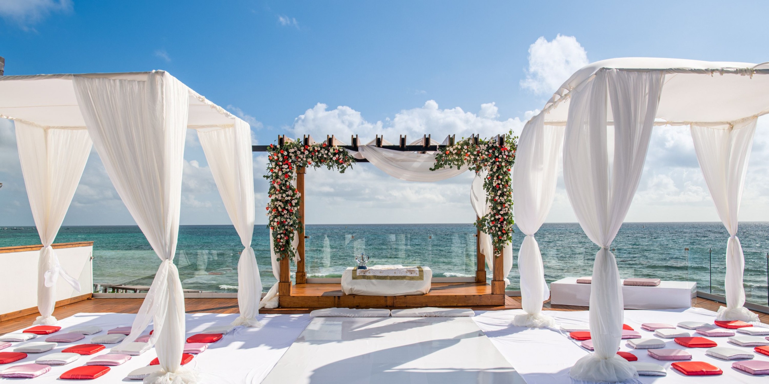 Experience a picturesque outdoor wedding at Hilton Playa del Carmen with an elegant altar featuring white canopy drapes and floral decorations, all set against the backdrop of the ocean under a clear blue sky.