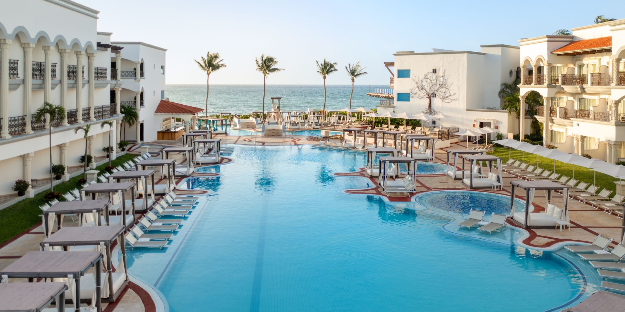 The large resort pool at the Hilton Playa del Carmen is surrounded by buildings and features loungers and cabanas. On a clear day, you can see the ocean and palm trees in the background.
