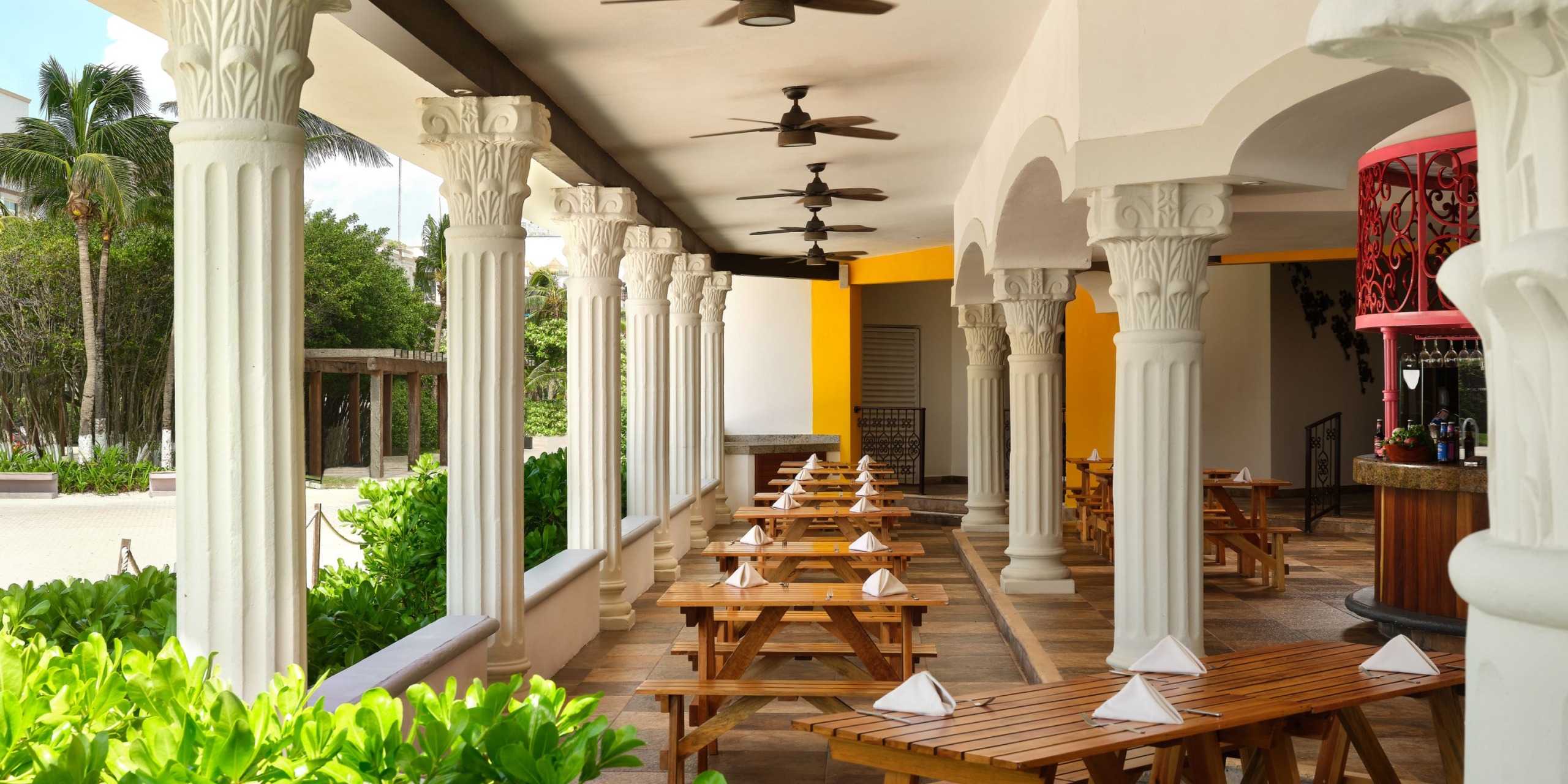 The Hilton Playa del Carmen offers an outdoor dining experience featuring wooden picnic tables complete with napkins, surrounded by elegant white columns and lush greenery, with ceiling fans providing comfort overhead.