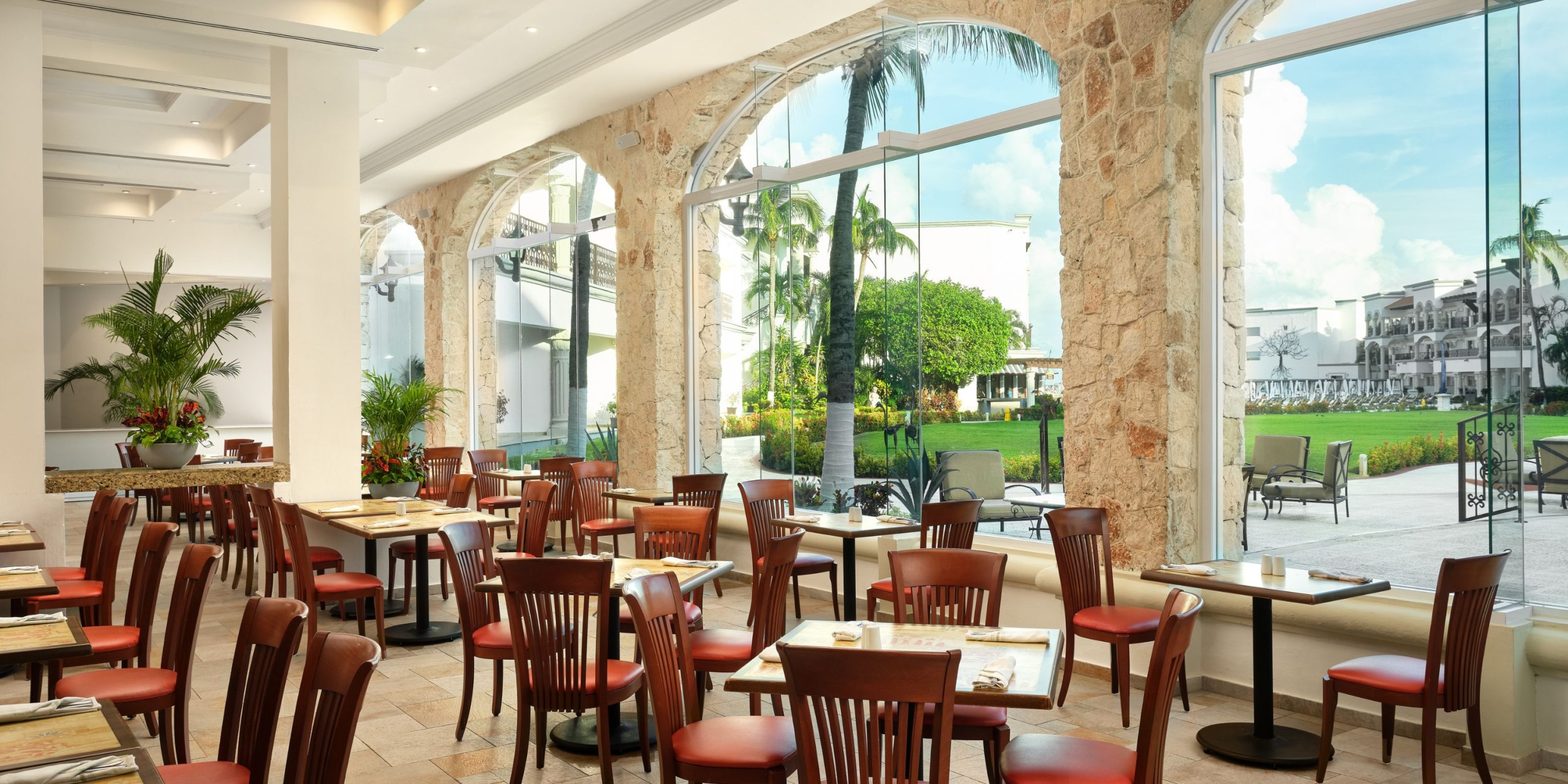 The Hilton Playa del Carmen features a bright restaurant interior adorned with wooden chairs and stone arches, offering views of a garden with palm trees through expansive windows. Dining tables are elegantly set for guests.