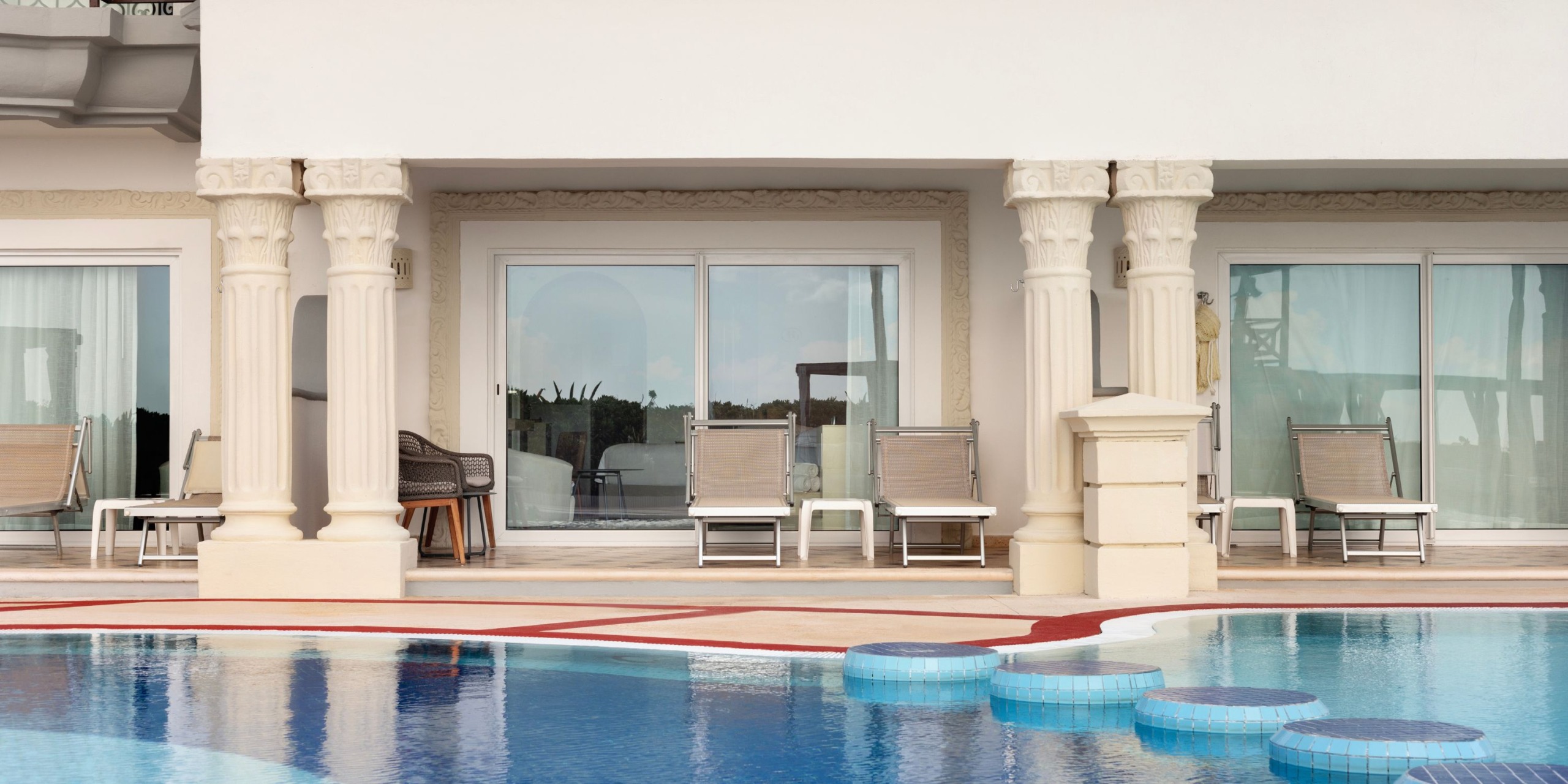 A luxurious poolside area at the Hilton Playa del Carmen features ornate columns, lounge chairs, and a sliding glass door leading into a building, with blue tiled steps offering access to the water.