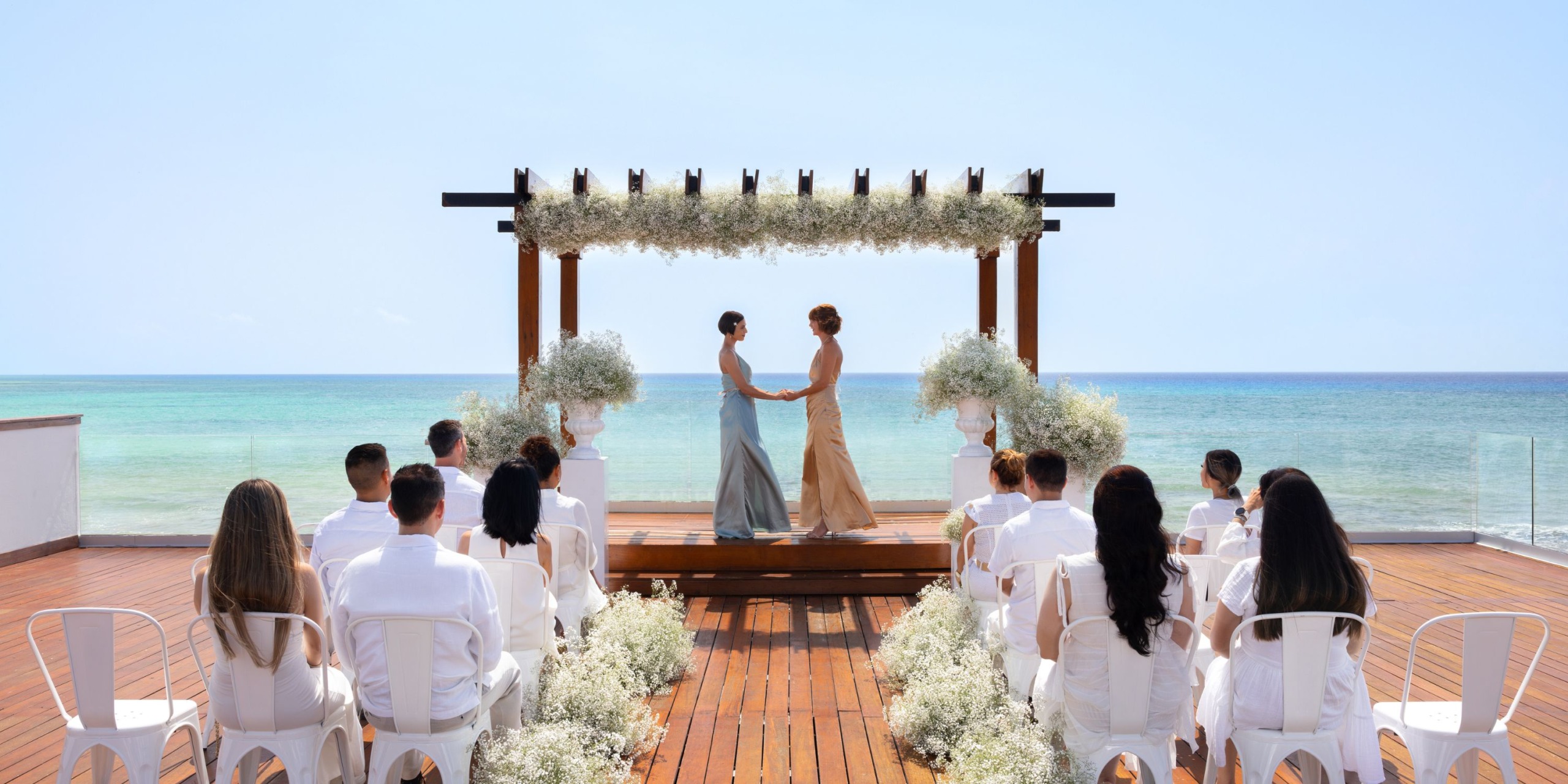Two people in formal attire stand under a floral arch by the ocean at the Hilton Playa del Carmen, holding hands, while guests sit on a wooden deck.