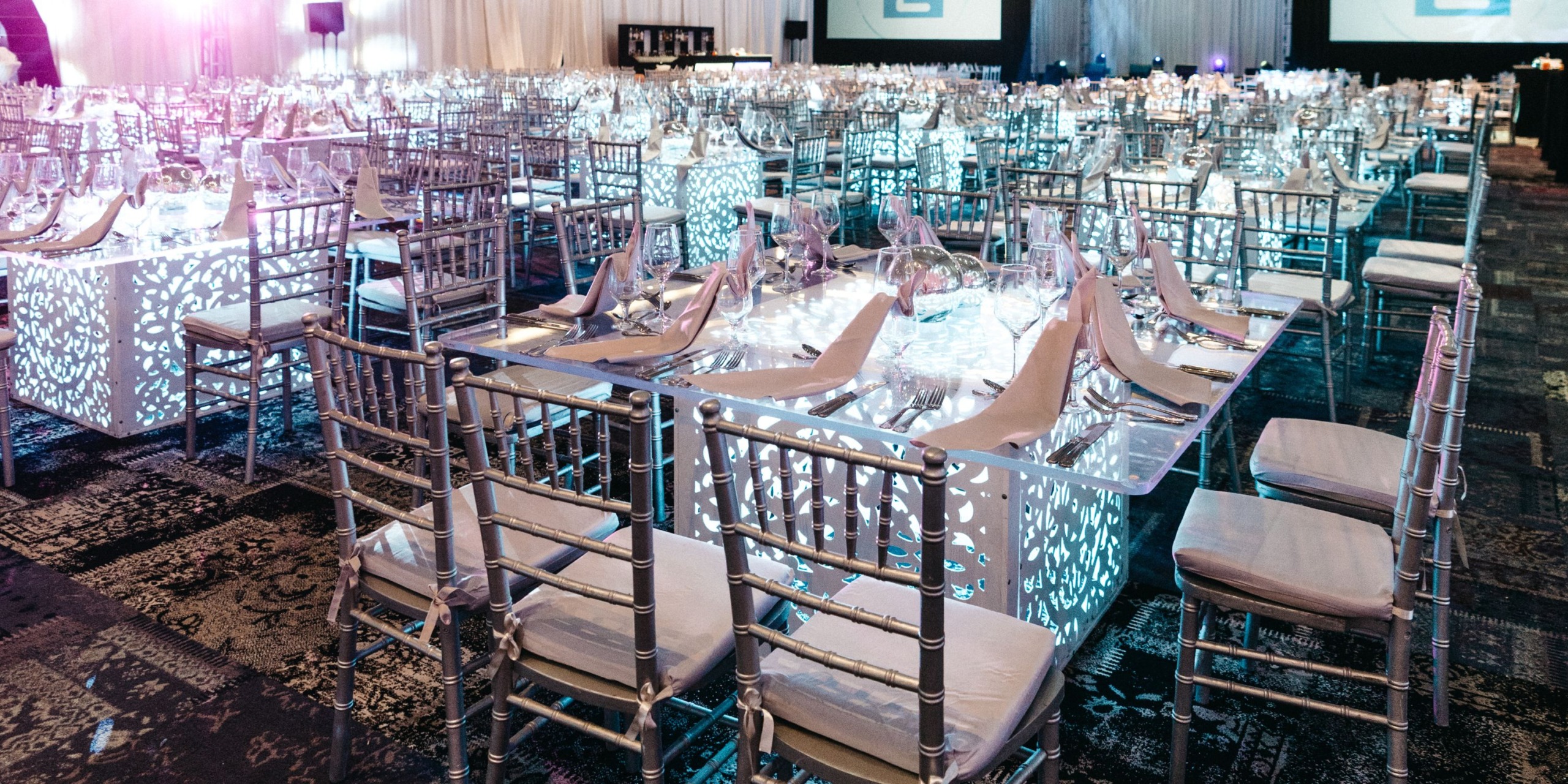 A formal event setup at Hard Rock Cancun features rows of tables with silver chairs in a spacious room. Tables are elegantly set with glasses and napkins, while the lighting creates a decorative pattern.