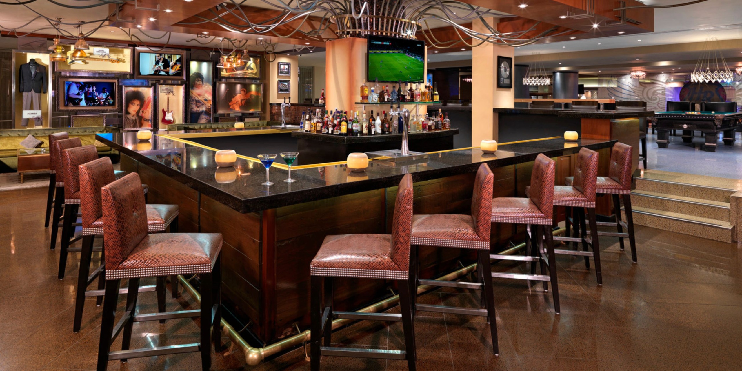 The Hard Rock Cancun offers a spacious bar with a U-shaped counter, complete with high chairs, an array of bottles, and a sports game playing on TV. The background is embellished with unique bar decor and additional seating options.