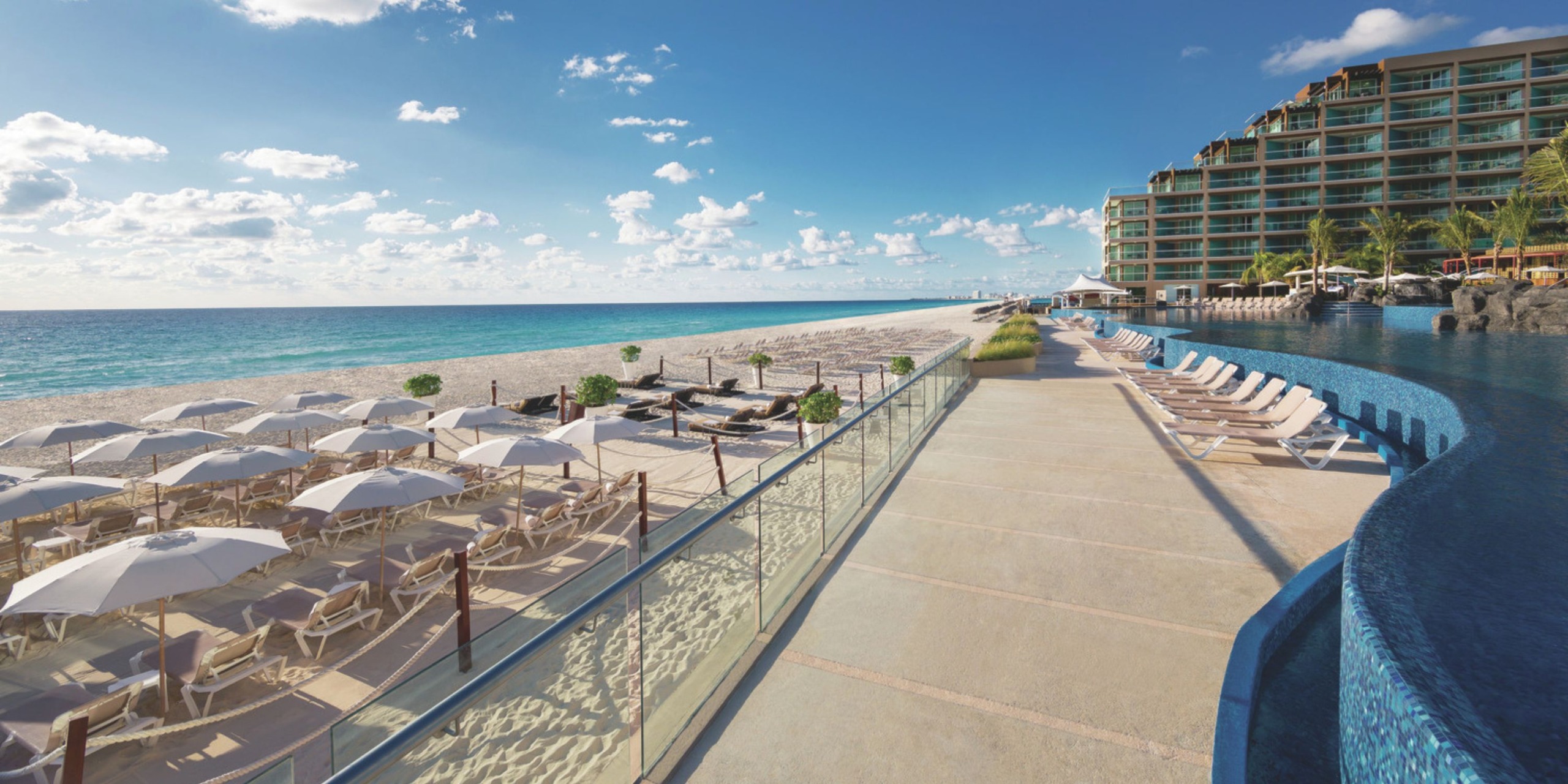 Hard Rock Cancun features a beachfront setting with a row of lounge chairs and umbrellas on the sand, an infinity pool to the right, and the hotel building in the background, all under a partly cloudy blue sky.