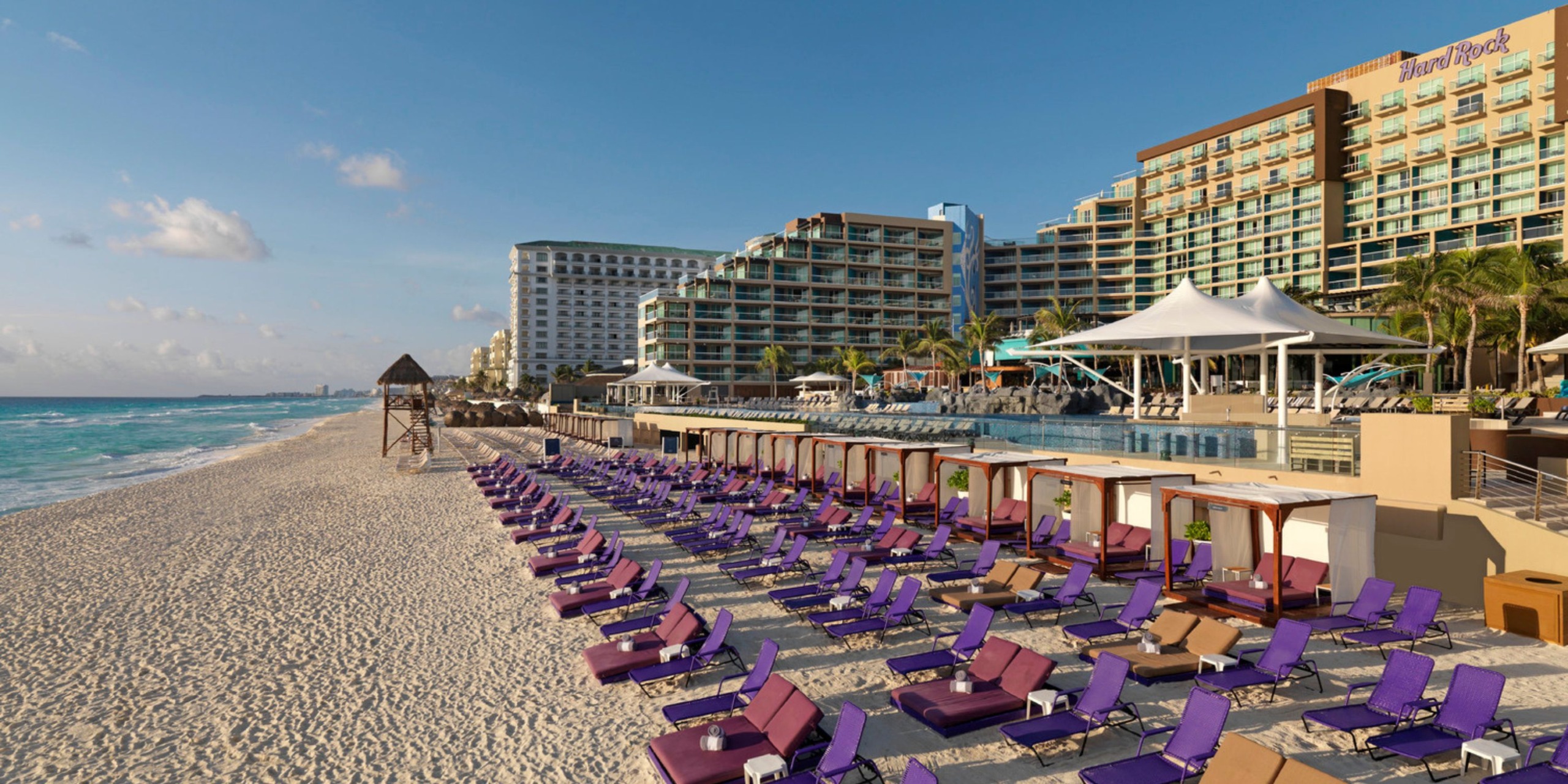 The Hard Rock Cancun boasts a stunning beachfront with purple lounge chairs perfectly arranged on the sandy shore. A lifeguard stand and the hotel buildings can be seen in the background beneath a clear blue sky.
