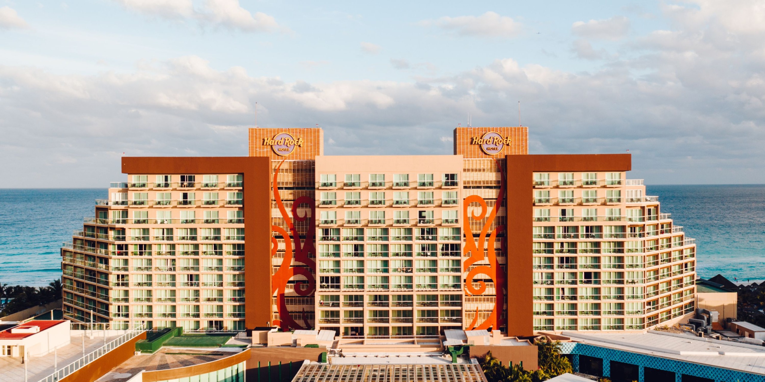 Aerial view of a large building with prominent "Hard Rock Cancun" signs on the facade, set against an ocean backdrop under a partly cloudy sky.