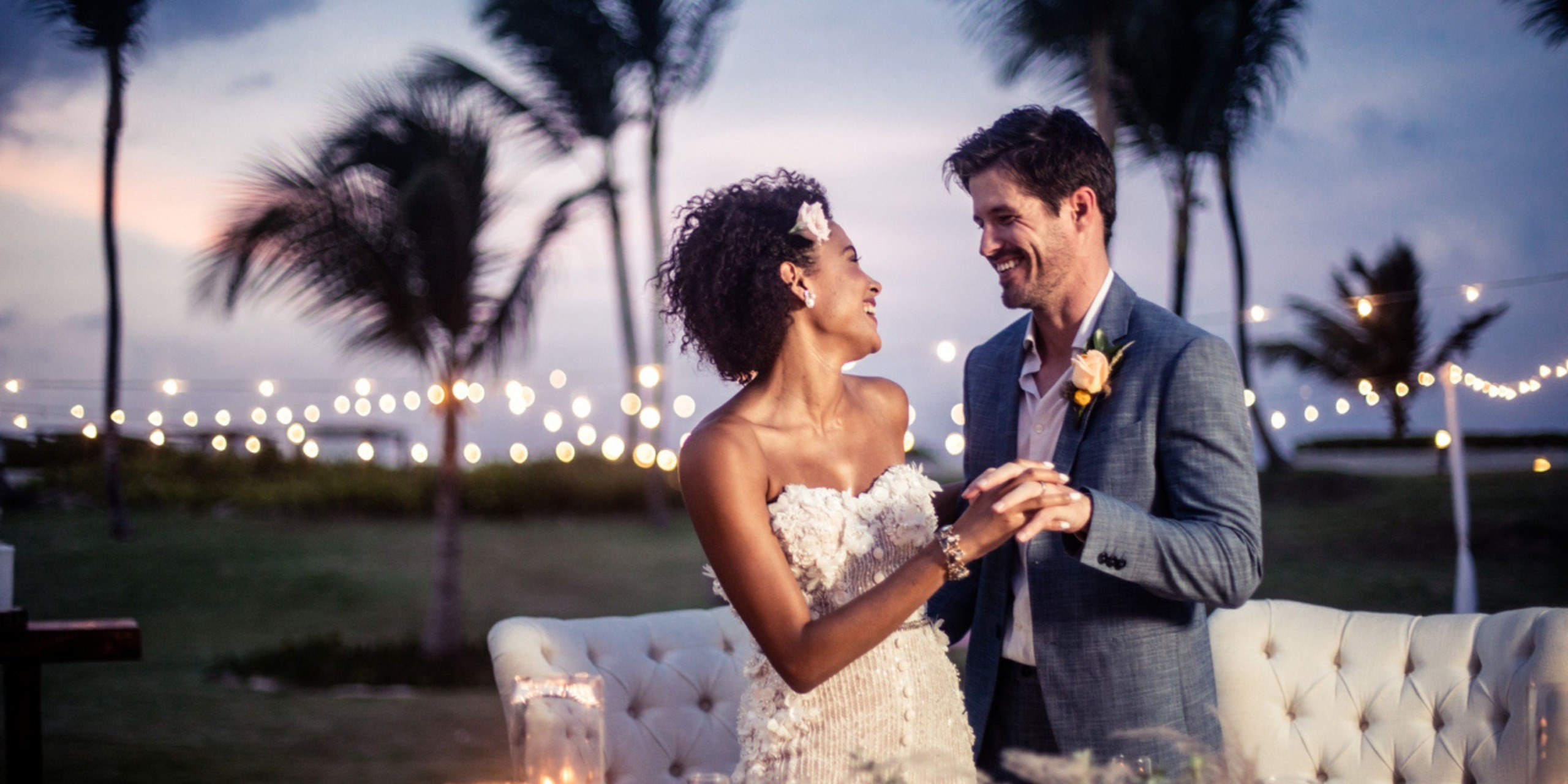 A couple in formal attire smiles and dances outdoors at dusk beside the enchanting backdrop of palm trees and string lights at Hard Rock Cancun.