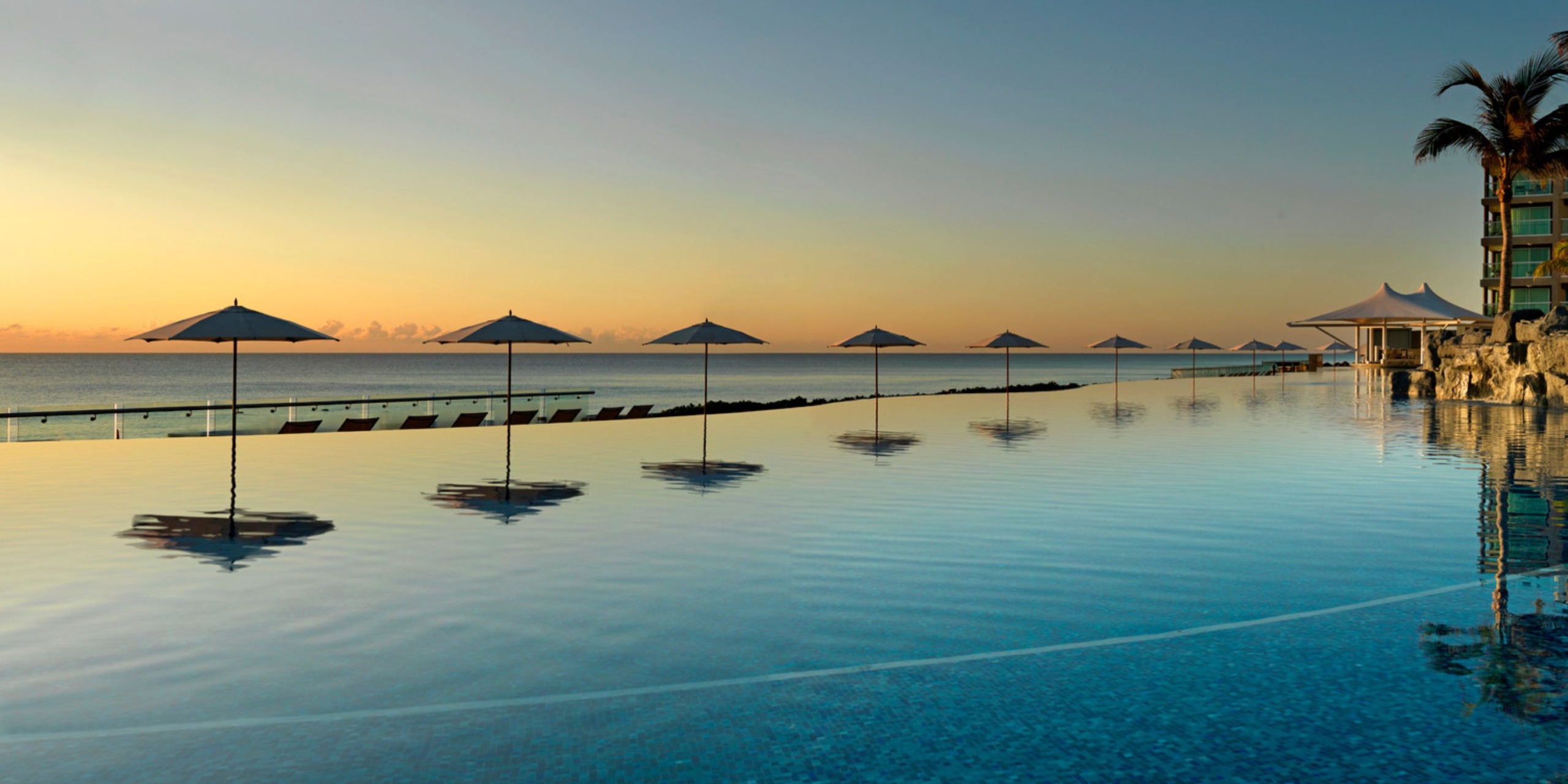 Gazing at the breathtaking sunset from the Hard Rock Cancun, guests can enjoy a tranquil infinity pool complete with a row of umbrellas and lounge chairs, all overlooking the vast ocean.