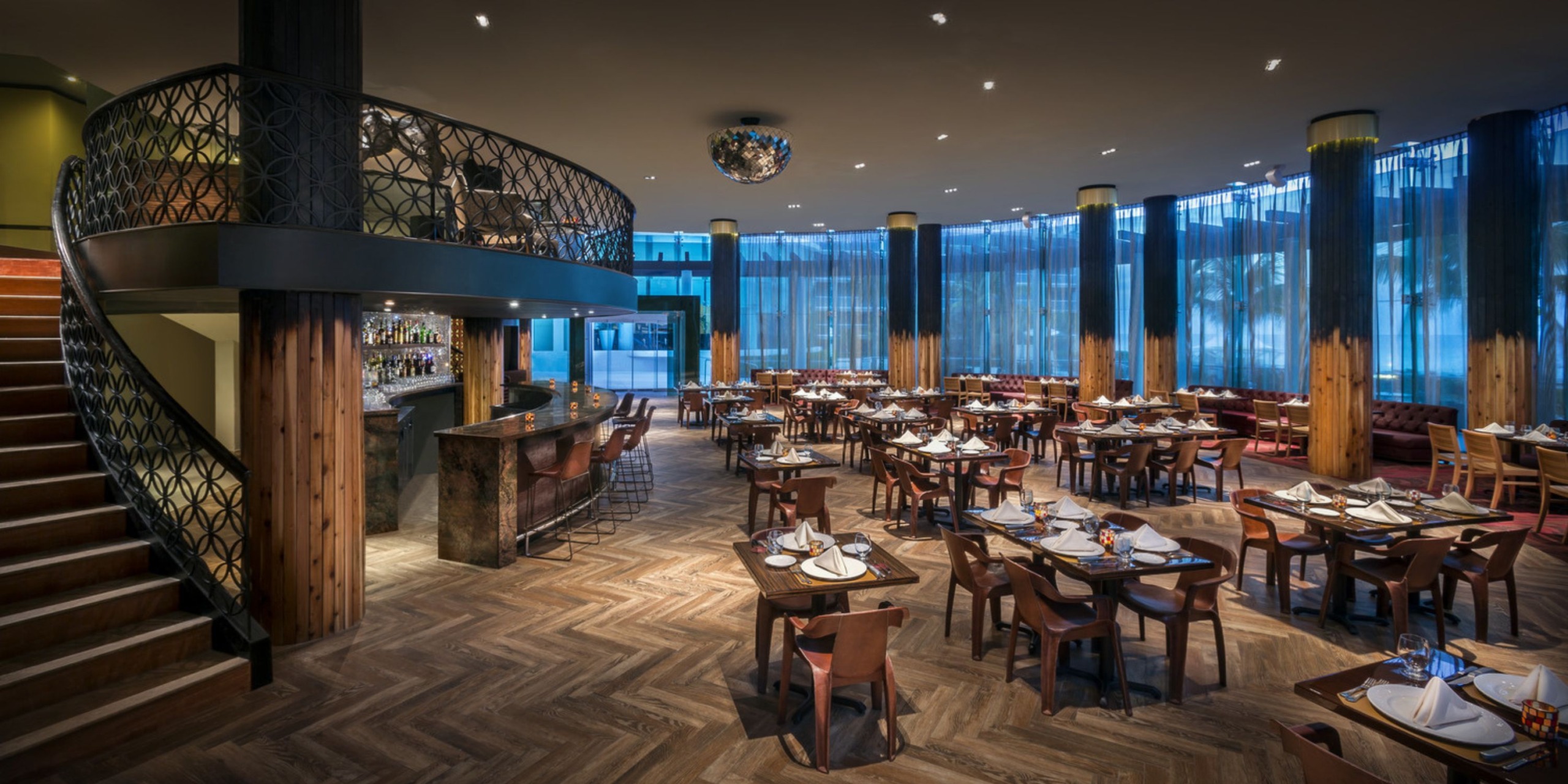 The Hard Rock Cancun boasts a contemporary dining space featuring wooden tables set for guests. A spiral staircase ascends to an upper level, while large windows adorned with blue curtains line the walls. On the left, patrons can enjoy the view of a well-appointed bar.