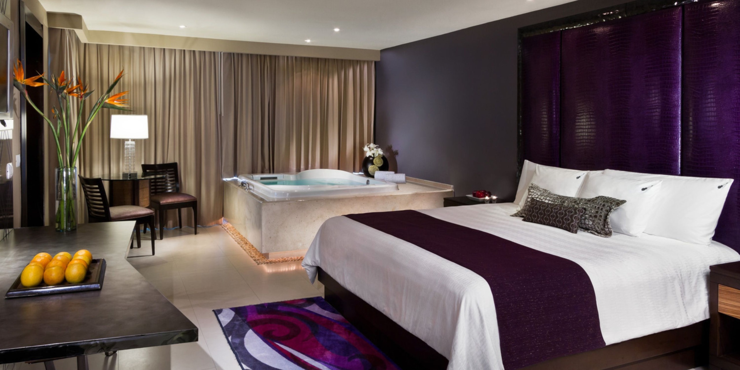 Experience the modern accommodations at Hard Rock Cancun, where you'll find a neatly made bed, a jacuzzi, and contemporary furniture accented with purple hues. The room also features a table topped with fresh fruit and a stylish lamp.