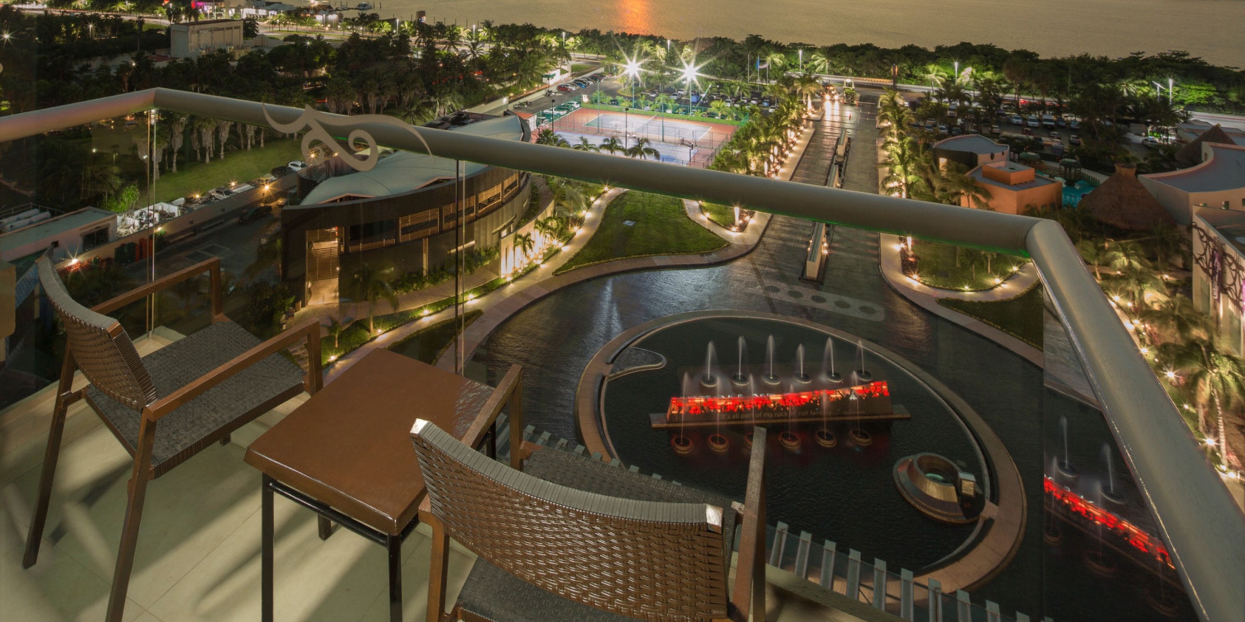 Balcony with chairs at the Hard Rock Cancun, offering views of a beautifully illuminated outdoor space that includes a fountain, bridge, and tennis court, all located near a waterfront setting at dusk.