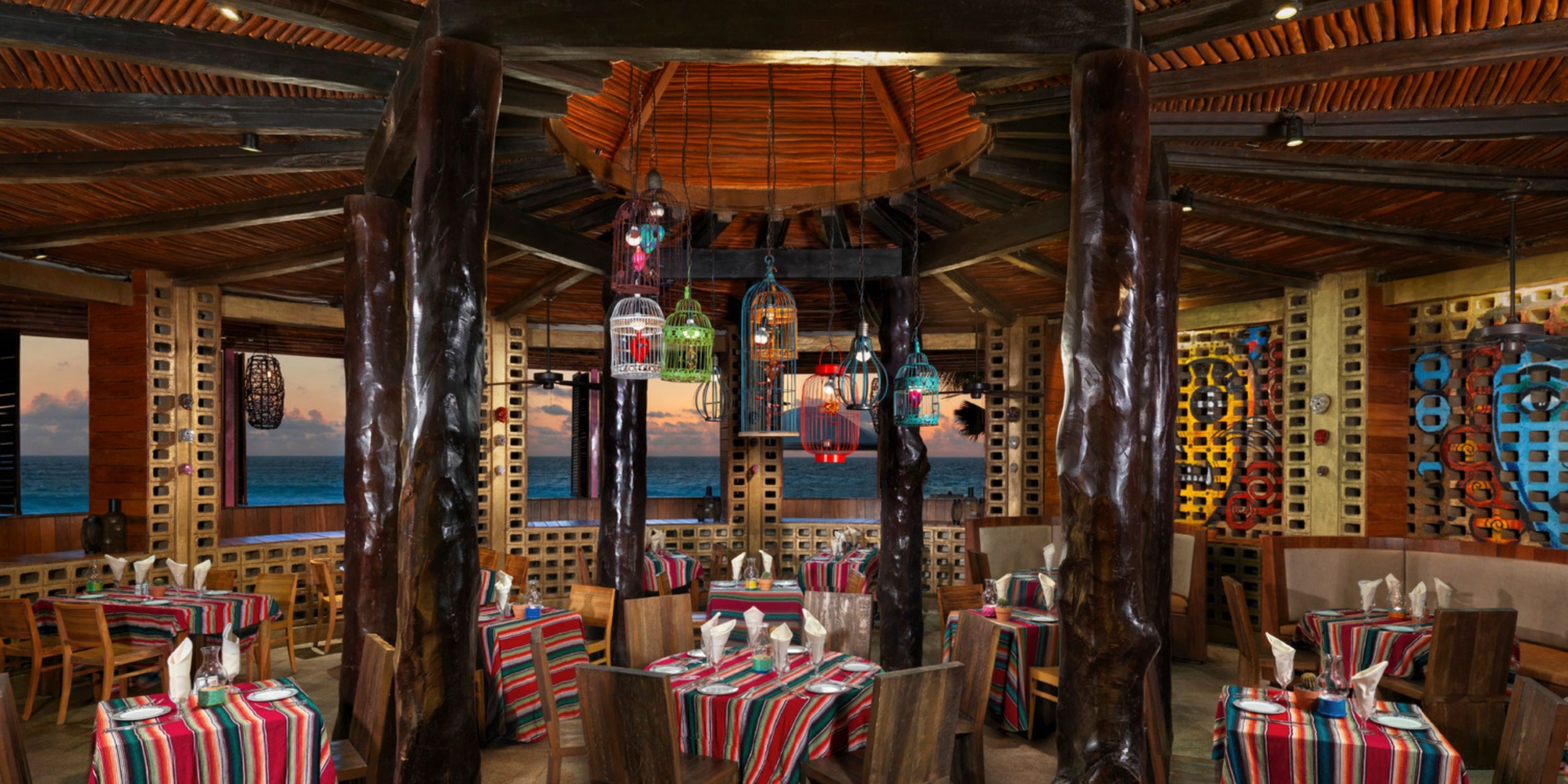 The Hard Rock Cancun boasts a rustic interior with wooden beams and vibrant tablecloths. Brightly colored lanterns hang from the ceiling, offering ocean views through large windows.
