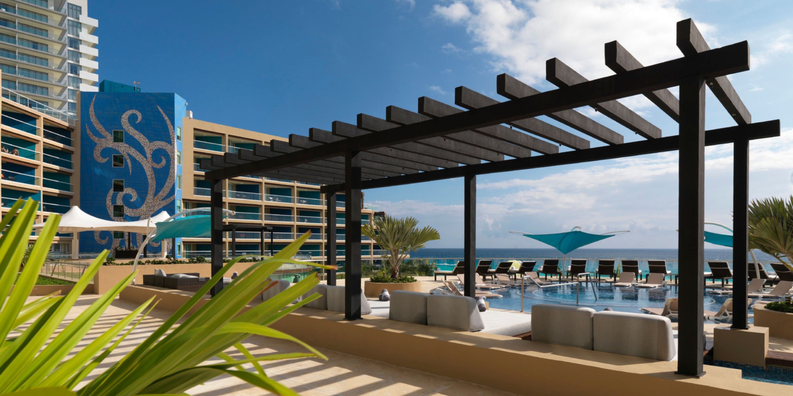 The Hard Rock Cancun features a contemporary outdoor pool area with sun loungers, a pergola, and stunning ocean views. It is surrounded by tall buildings, with blue sails providing ample shade.