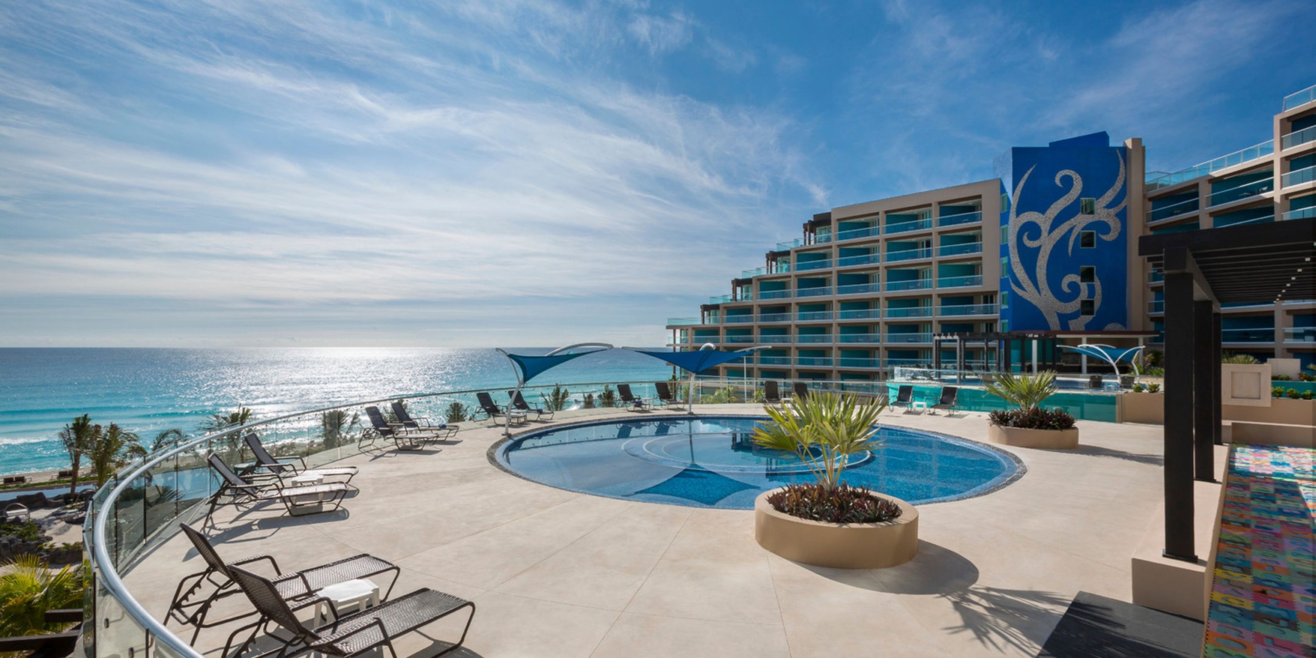 A rooftop pool area with lounge chairs at the Hard Rock Cancun offers stunning ocean views, while a modern building with large windows stands in the background under a clear blue sky.