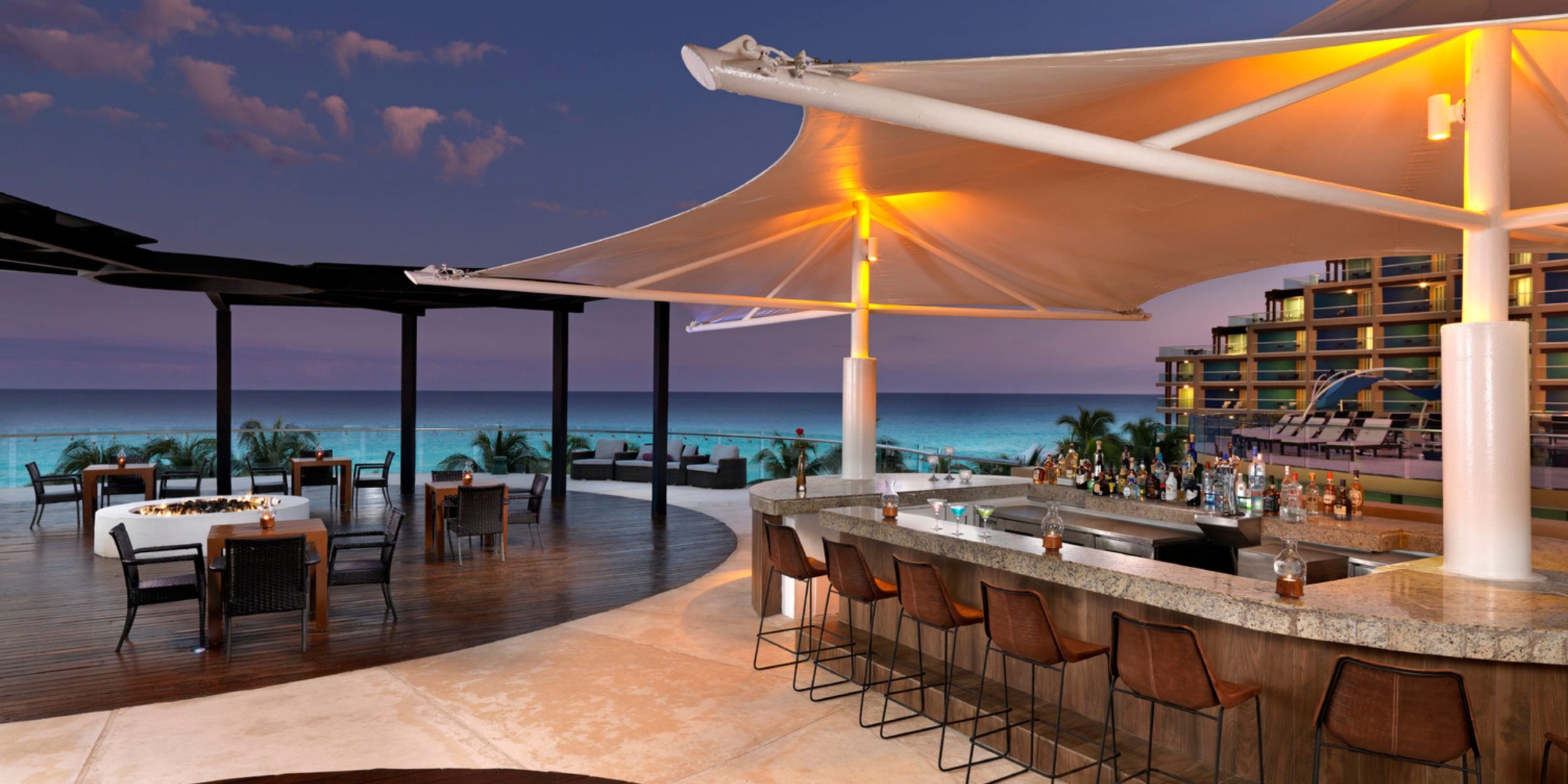 The Hard Rock Cancun features a contemporary outdoor bar with a gracefully curved canopy, offering stunning ocean views. Guests can relax on the wooden deck, furnished with tables and chairs, while enjoying vistas of the beach and horizon.