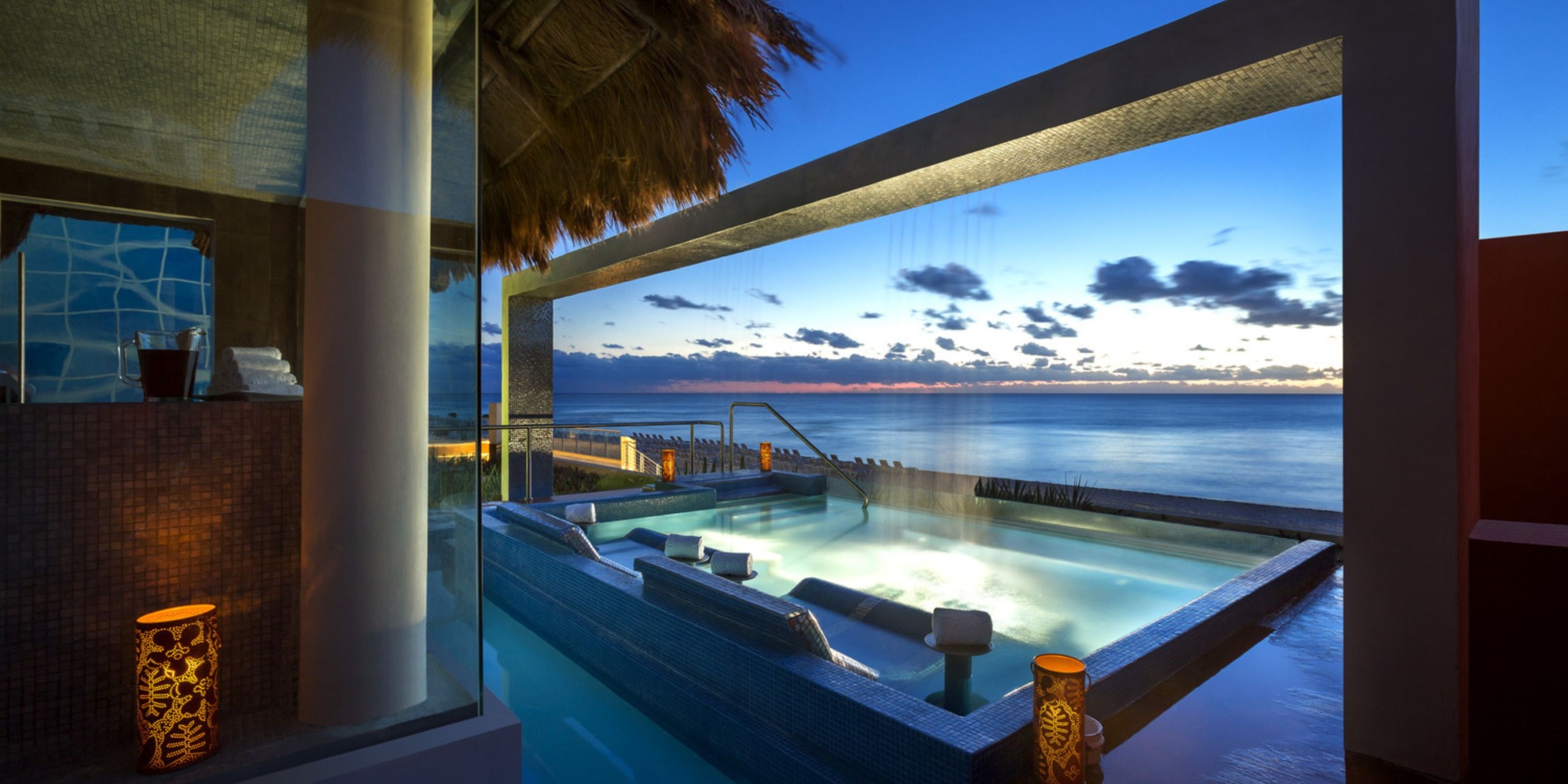 Experience an outdoor infinity pool at Hard Rock Cancun, featuring submerged lounge chairs that offer breathtaking views of the ocean at sunset. The ambient atmosphere is enhanced by lit lanterns and a cascading waterfall.