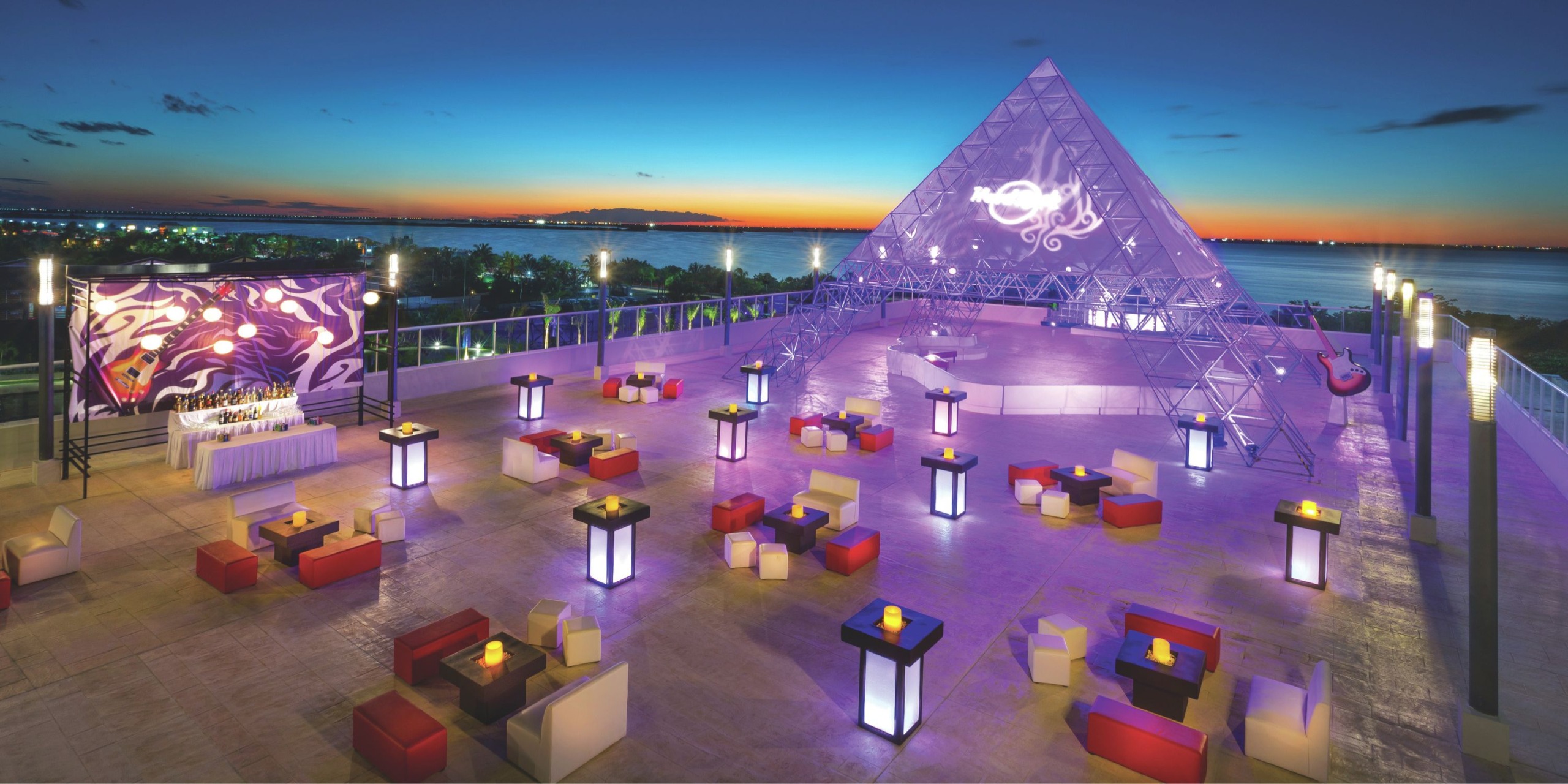 A rooftop venue at Hard Rock Cancun offers modern seating, illuminated tables, and a large pyramid structure that overlooks a scenic sunset view of the water and distant land.