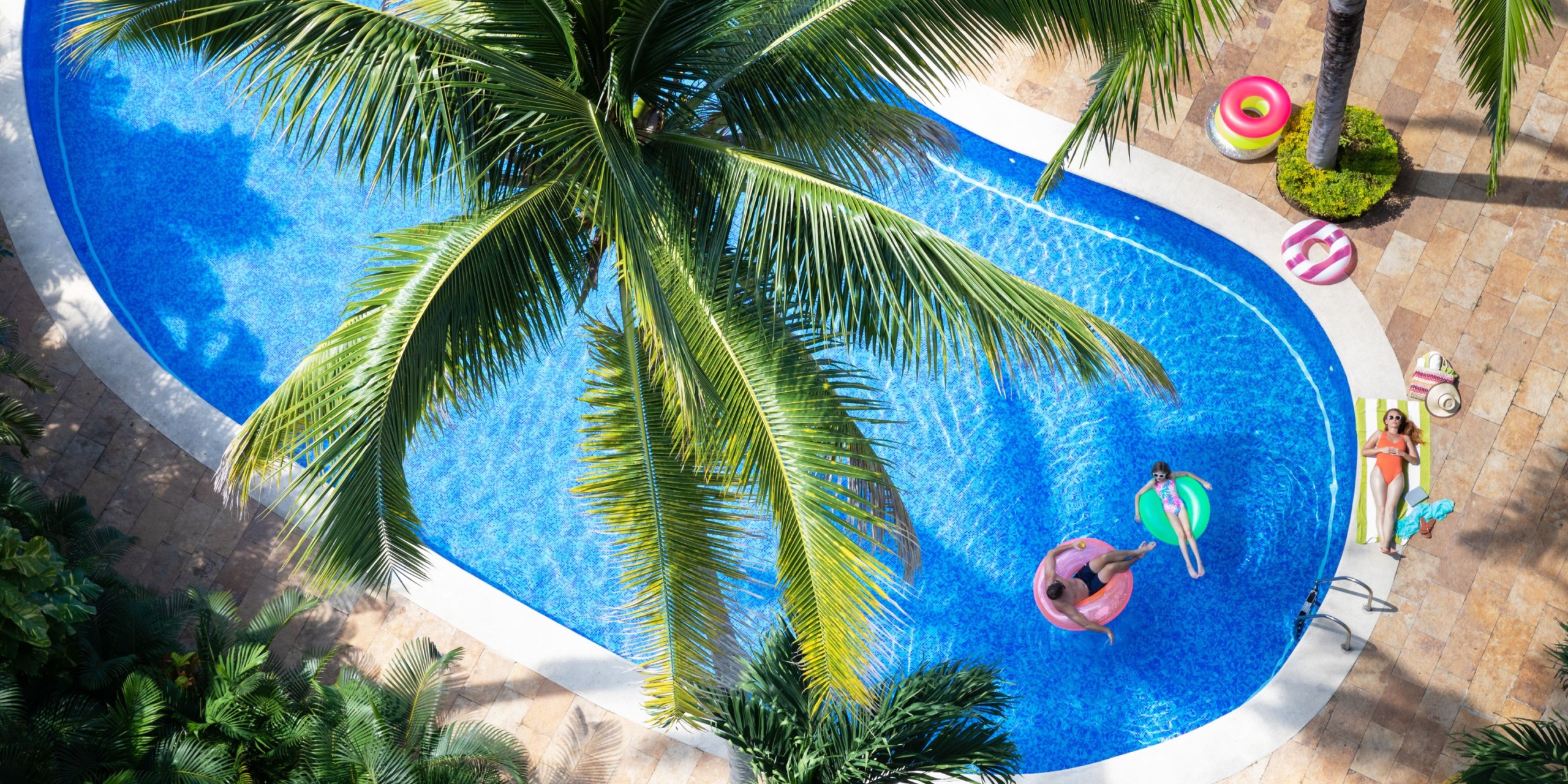 The Hyatt Ziva Puerto Vallarta offers an aerial view of its curvy swimming pool, beautifully surrounded by palm trees. Guests enjoy the water on inflatable rings, while others relax on sunbeds nearby. The scene is completed with a scattering of colorful pool floats around the area.