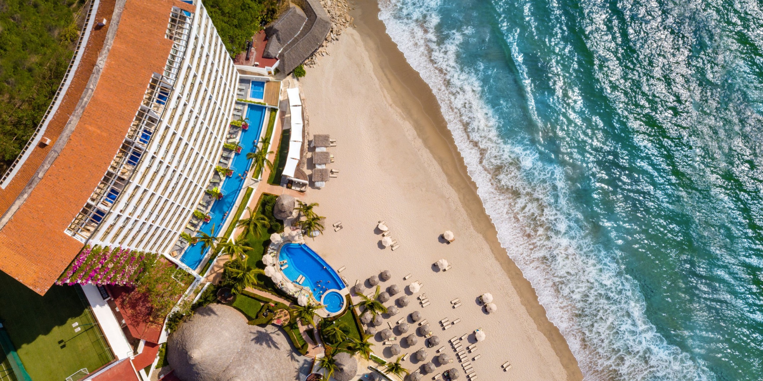 Aerial view of the Hyatt Ziva Puerto Vallarta, a beachfront resort featuring a large building, two pools, palm trees, and umbrellas on the sandy beach next to the ocean.