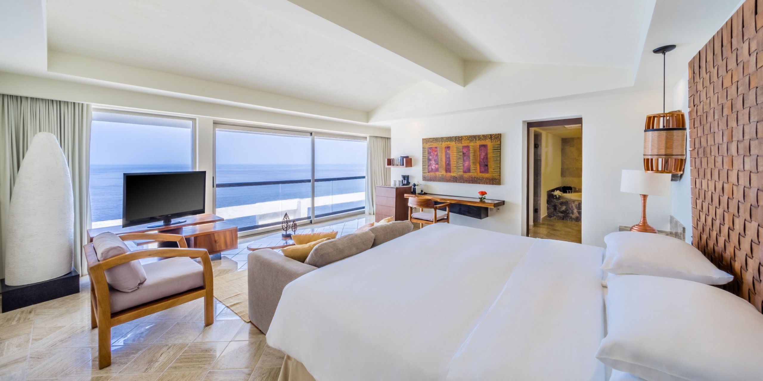 Spacious Hyatt Ziva Puerto Vallarta room with ocean view, offering a bed, armchair, TV, desk, and expansive windows. The walls showcase light and wooden tones adorned with framed artwork and complemented by a decorative lamp.