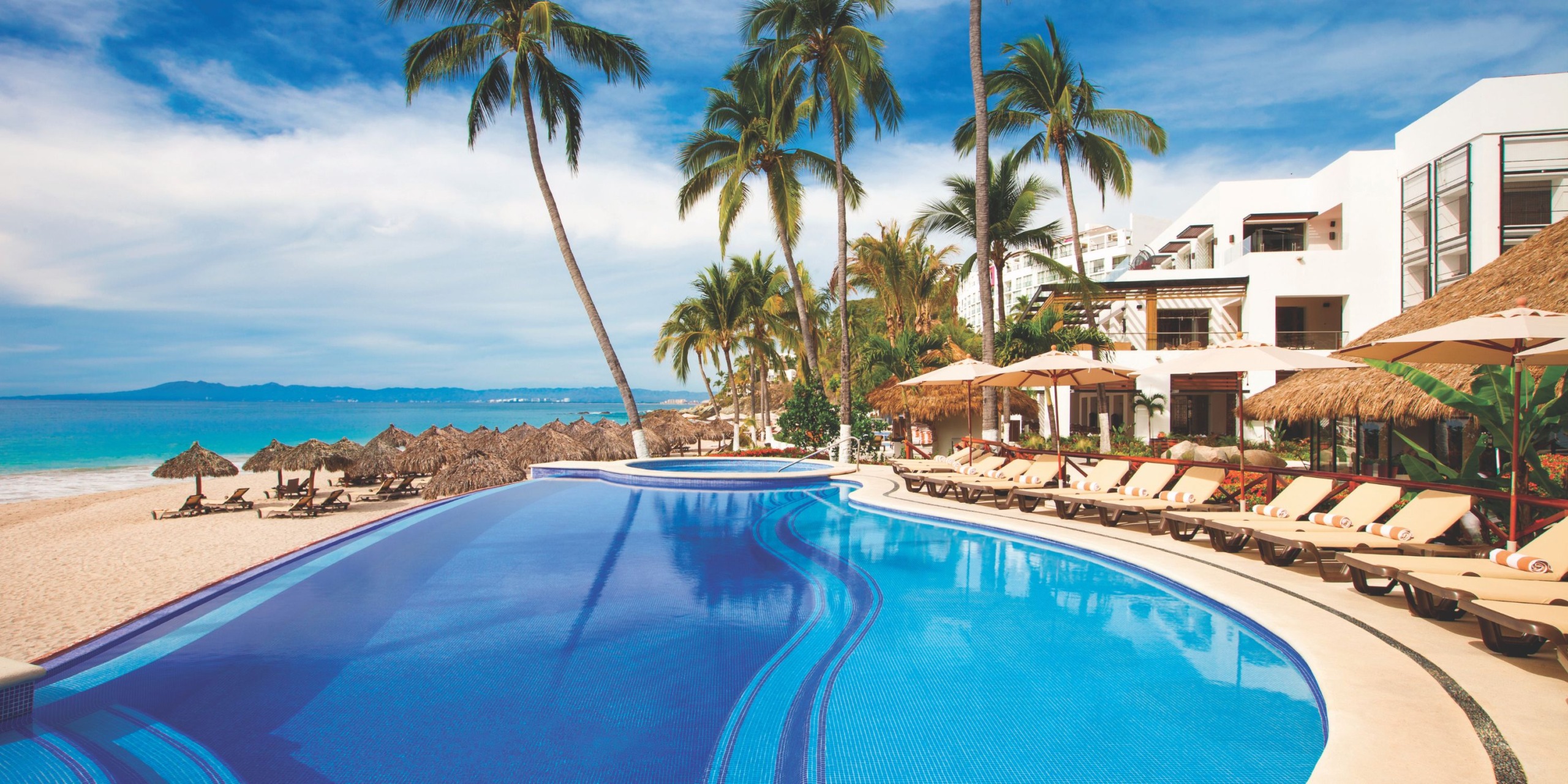 Experience the Hyatt Ziva Puerto Vallarta, a beachfront resort featuring an infinity pool encircled by lounge chairs and palm trees, complemented by thatched umbrellas on the sandy beach under a clear blue sky.