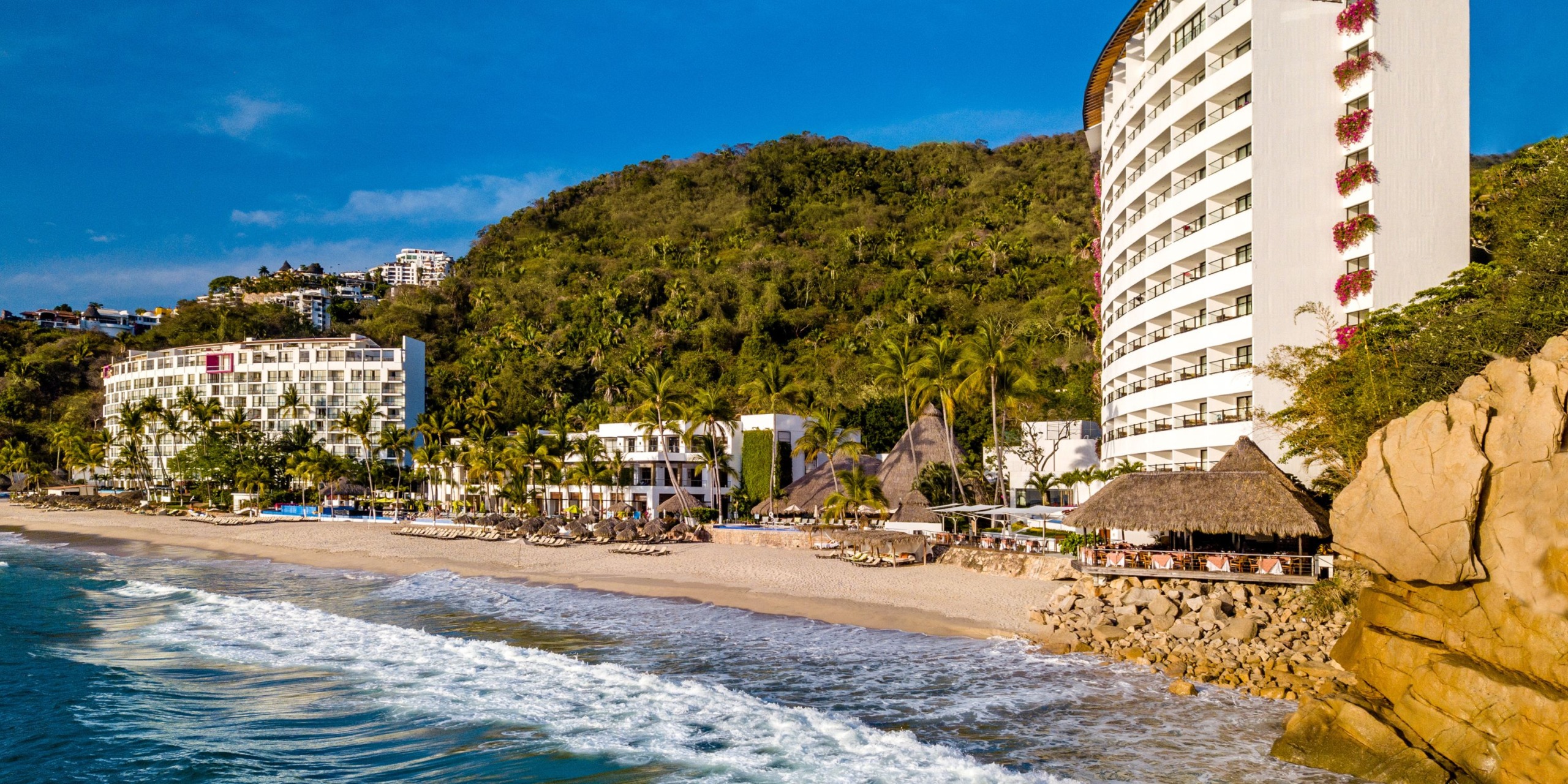 Hyatt Ziva Puerto Vallarta, a beachfront resort, features white high-rise buildings nestled among palm trees and a charming thatched roof structure. The sandy shore meets gently lapping waves, framed by lush green hills rising in the background under a blue sky.