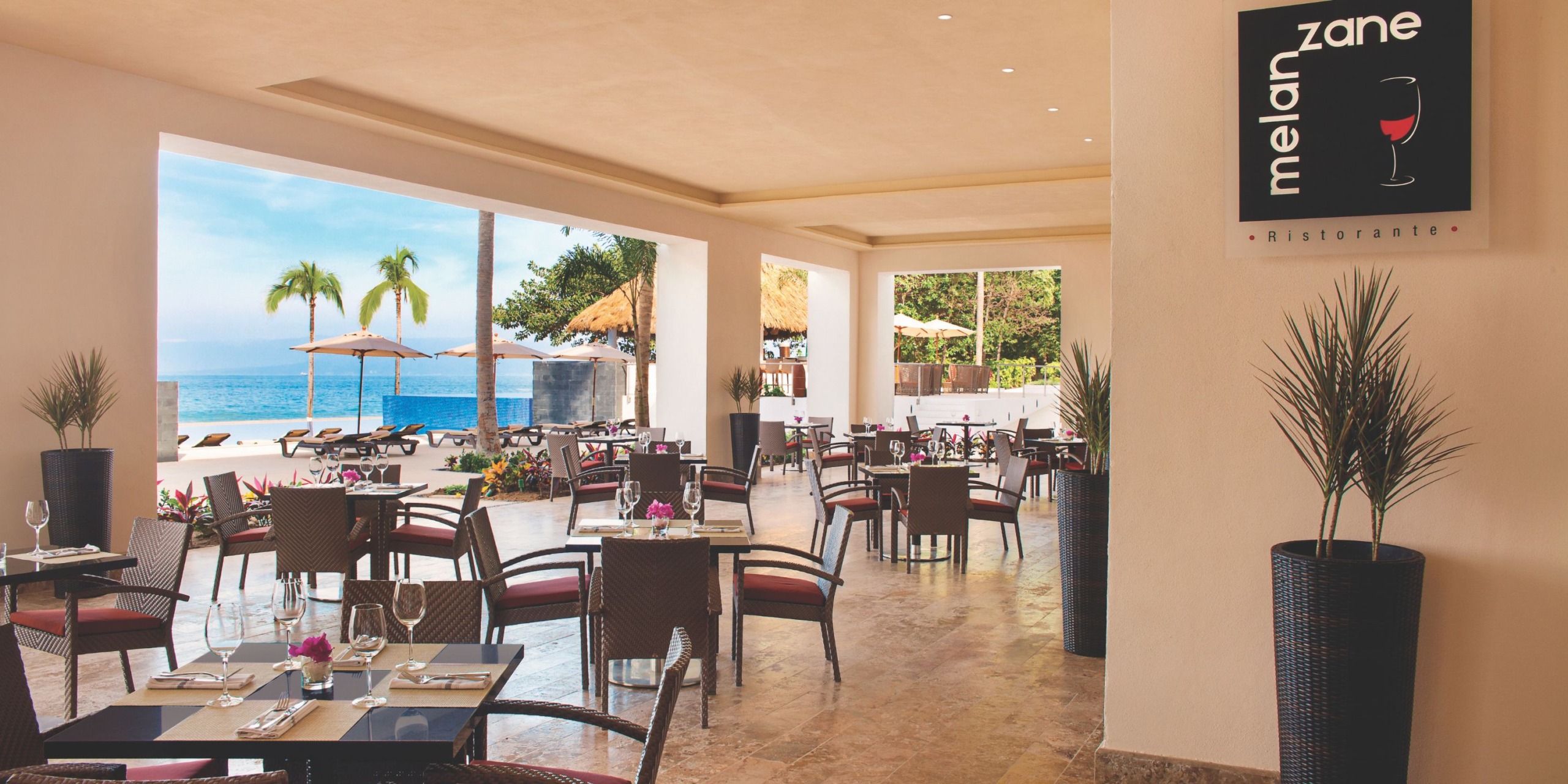 An outdoor restaurant featuring wicker chairs and tables, offering a breathtaking view of the beach. Surrounded by palm trees with a clear blue sky above, a sign displays "melanzane Ristorante" at Hyatt Ziva Puerto Vallarta.