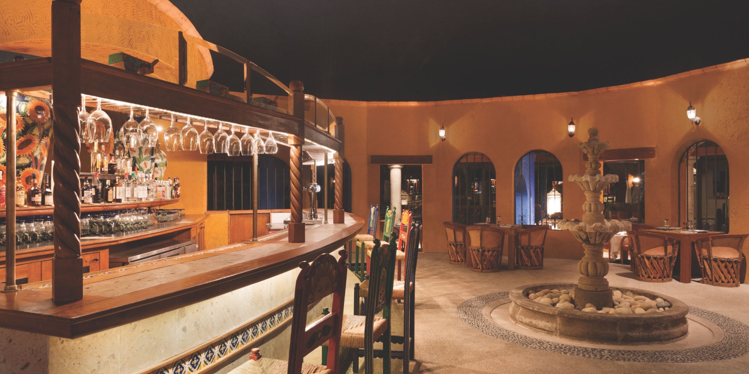 The interior of Hyatt Ziva Puerto Vallarta features a rustic bar and restaurant with wooden furniture, a circular bar, a hanging glass rack, a decorative fountain, and ambient lighting.