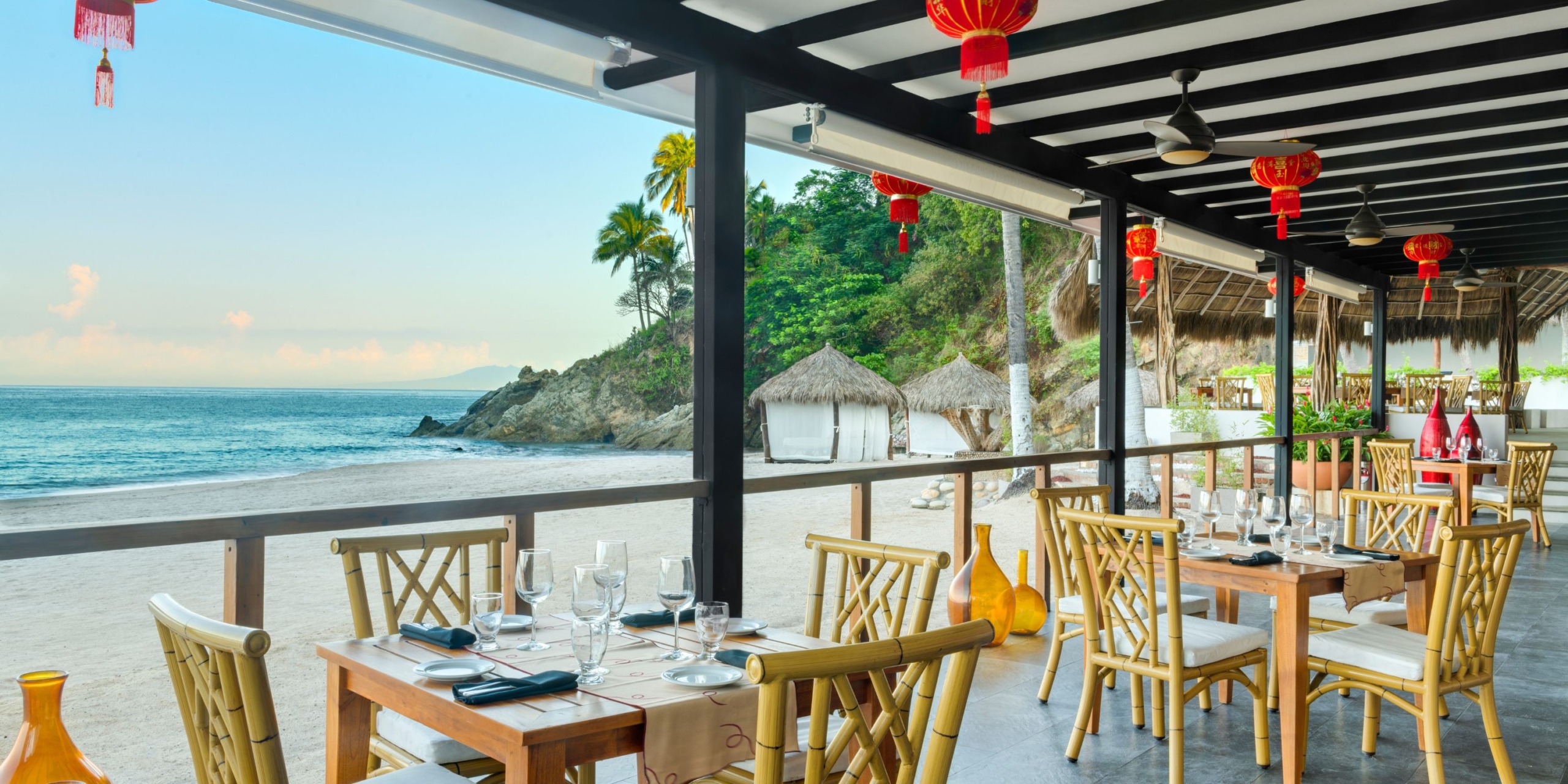 The beachfront restaurant at Hyatt Ziva Puerto Vallarta features wooden tables and chairs, embellished with red lanterns. Guests can enjoy stunning views of the ocean, sand, and trees in the background.