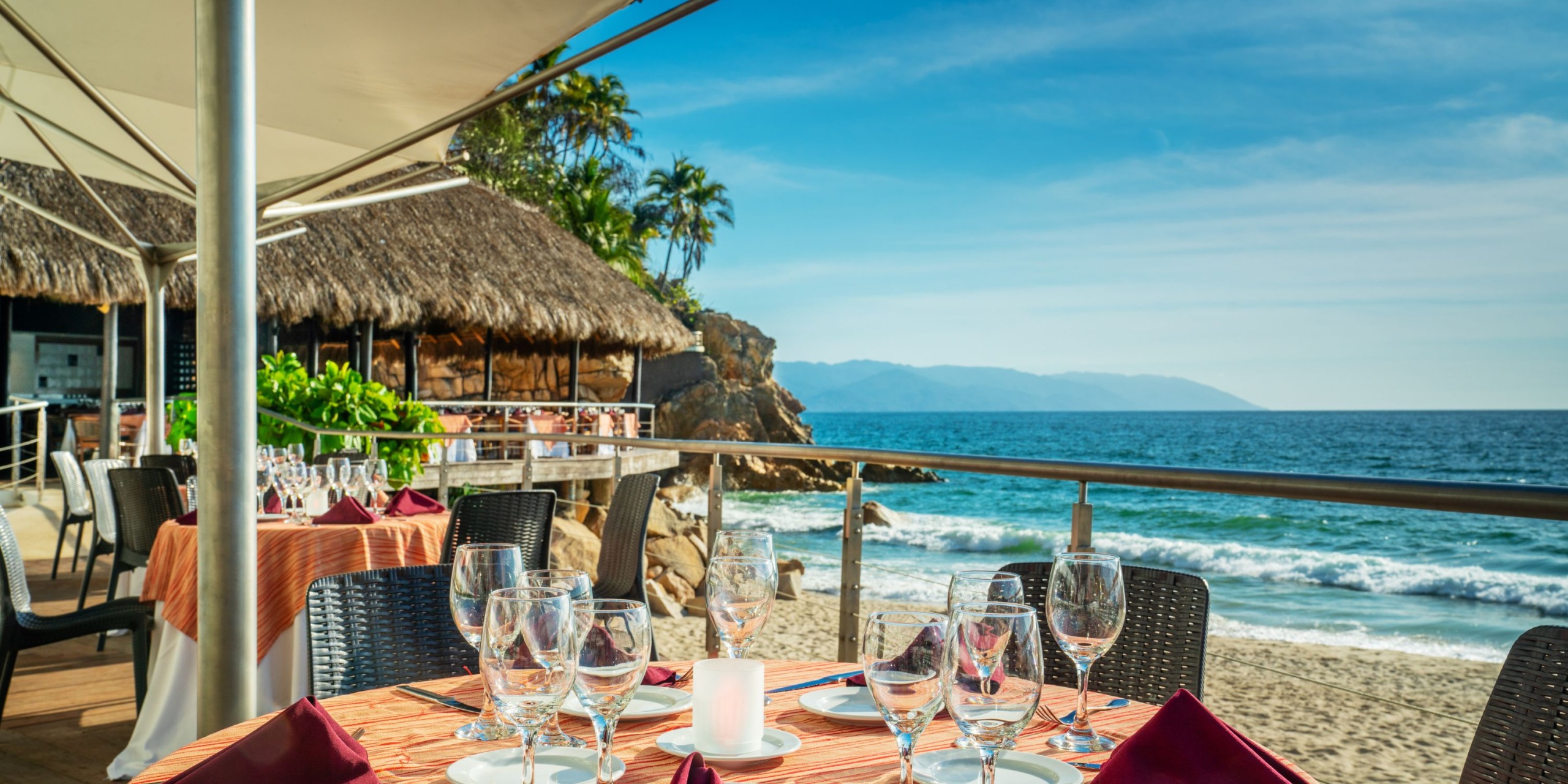 Enjoy a meal at the oceanfront Hyatt Ziva Puerto Vallarta, where tables are arranged under a canopy. You'll see a thatch-roofed hut in view, framed by the backdrop of waves and mountains.