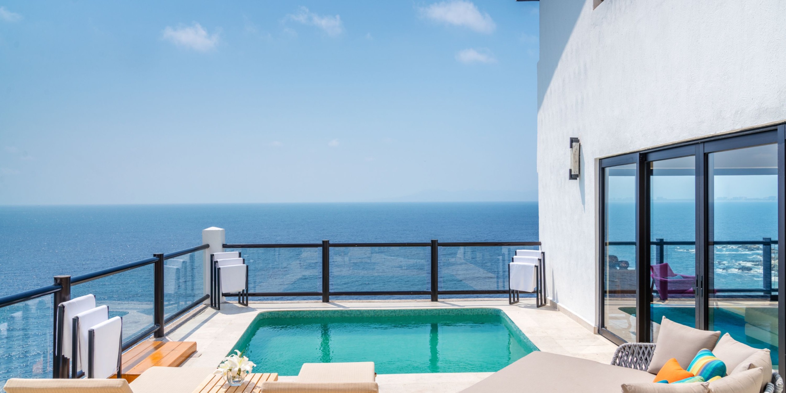 The Hyatt Ziva Puerto Vallarta features an infinity pool on a terrace with lounge chairs and glass railings, all overlooking the ocean under a blue sky.
