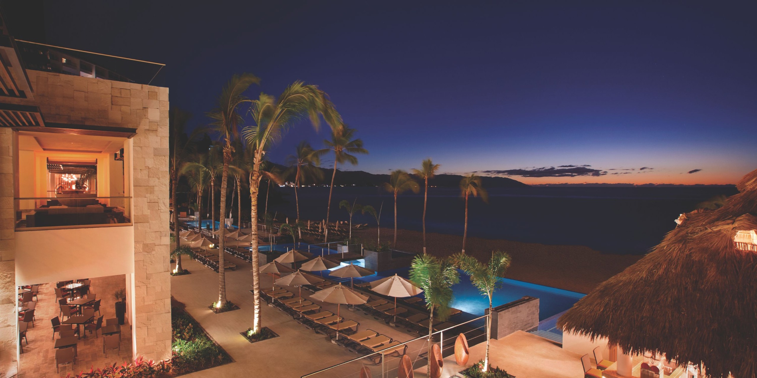 Hyatt Ziva Puerto Vallarta at dusk features palm trees, lounge chairs, umbrellas, and a beautifully illuminated pool. The building emits a warm glow while the horizon showcases a colorful sunset sky.