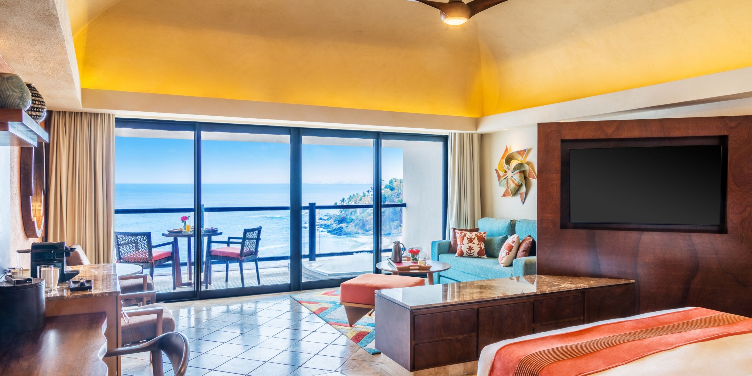 Experience a stay at the Hyatt Ziva Puerto Vallarta, offering a luxurious room with a large bed, comfortable seating area, and a private balcony. The balcony is furnished with a table and chairs, providing breathtaking ocean views. Enjoy the warm lighting throughout the room and entertain yourself with the flat-screen TV.