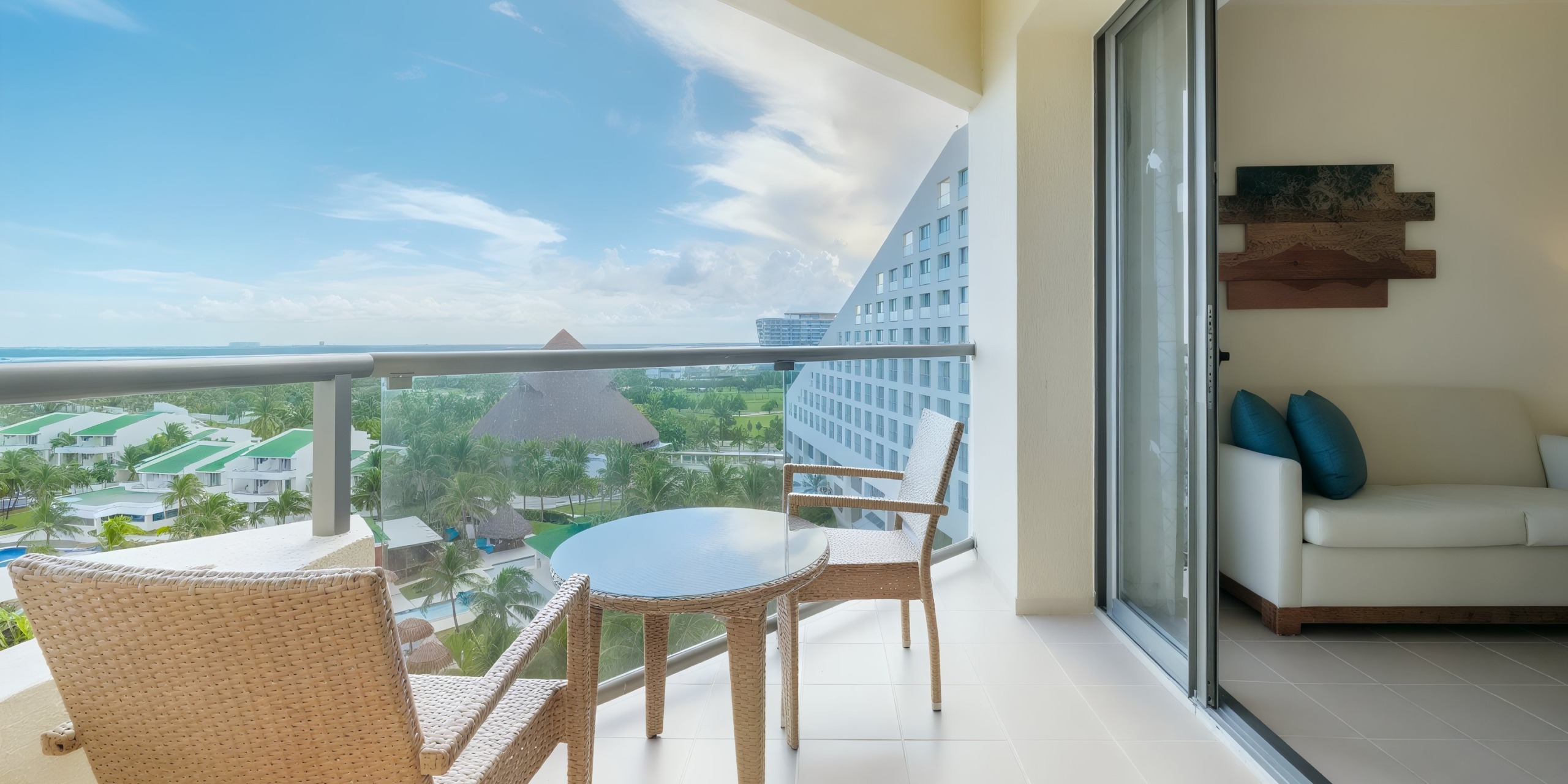 Balcony with two wicker chairs and a round table overlooking Iberostar Selection Cancun's resort area featuring a pool and lush greenery; a modern building is visible on the right, with a partially open sliding glass door.
