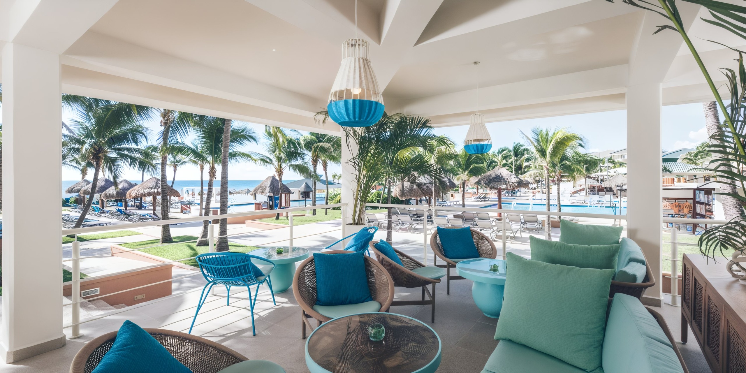 Experience the Iberostar Selection Cancun's outdoor patio with turquoise seating and tropical decor, offering stunning views of a pool and beach. Palm trees and thatched huts provide a picturesque backdrop.