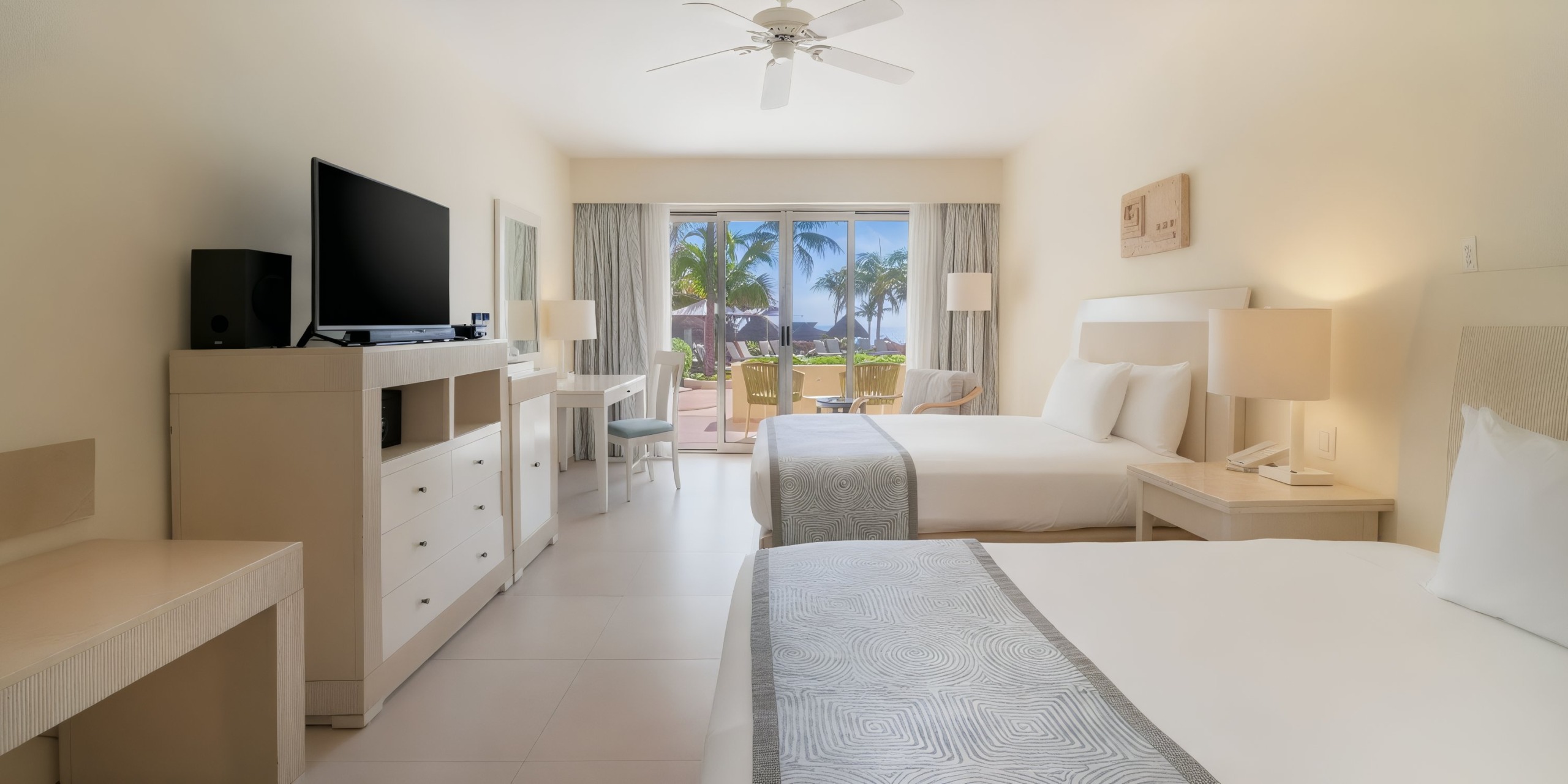 Experience the Iberostar Selection Cancun, featuring a hotel room with two beds and a TV on a dresser under a ceiling fan. Enjoy the breathtaking view of palm trees and the ocean through a large window, all set in modern and minimalist decor.