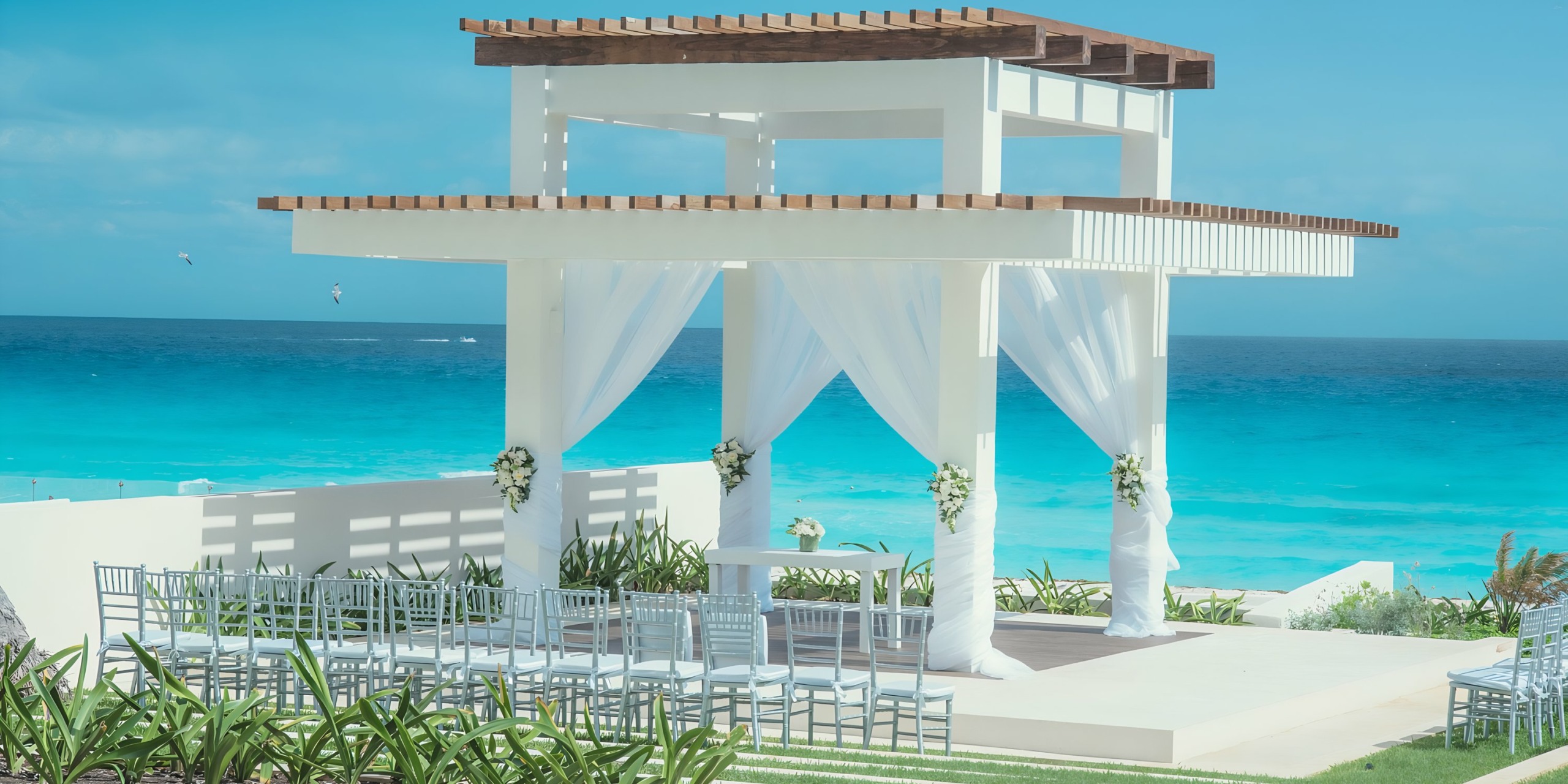 An Iberostar Selection Cancun oceanfront wedding gazebo setup with white drapery and floral decorations under a clear blue sky.