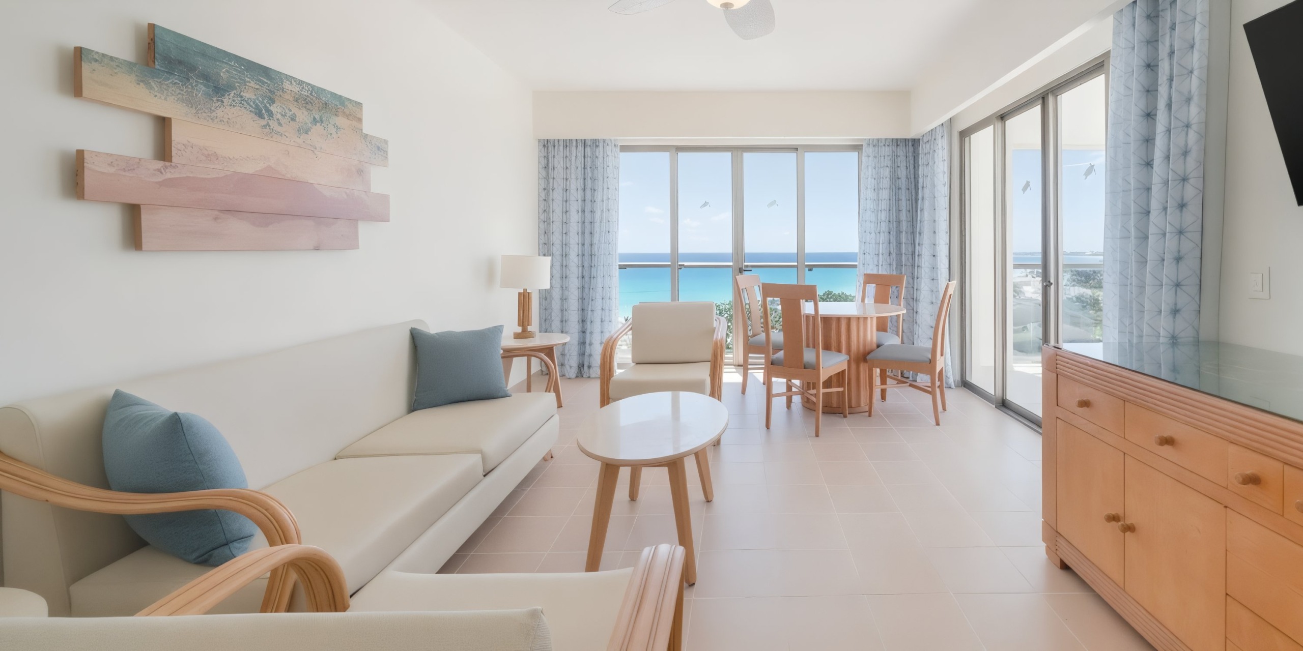 A bright living room at the Iberostar Selection Cancun features beige furniture, a wooden coffee table, and a dining set by the window. Large windows provide an ocean view, with coastal artwork adorning the walls.