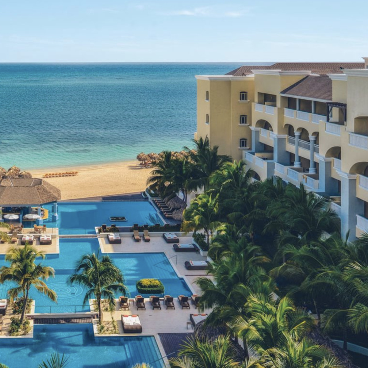 Iberostar Grand Hotel Rose Hall perfect for destination weddings, with a large swimming pool overlooking the ocean, surrounded by palm trees under a clear sky.