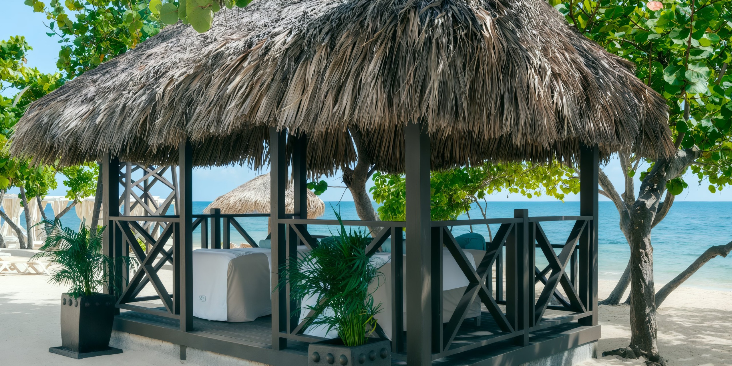 The Iberostar Waves Rose Hall Beach features a charming thatched-roof cabana with lounge chairs set amidst trees on a sandy beach, offering serene views of the calm ocean.