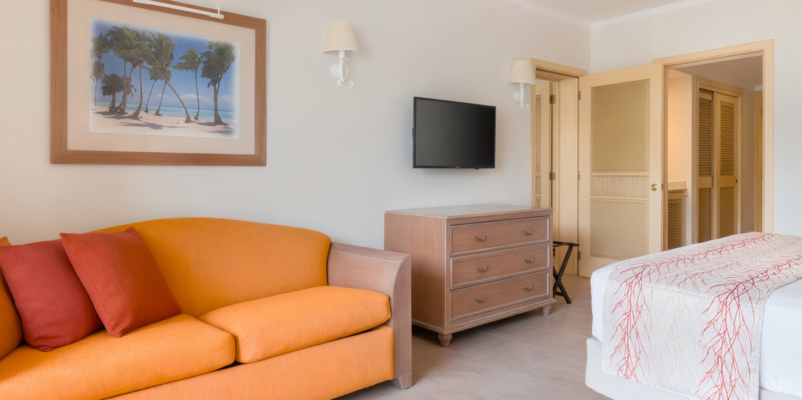 Experience the Iberostar Waves Rose Hall Beach, featuring a hotel room adorned with an orange couch, a wall-mounted TV, a dresser, and an open closet. The room includes a bed with elegant white and red bedding and is accented by a framed beach picture on the wall.