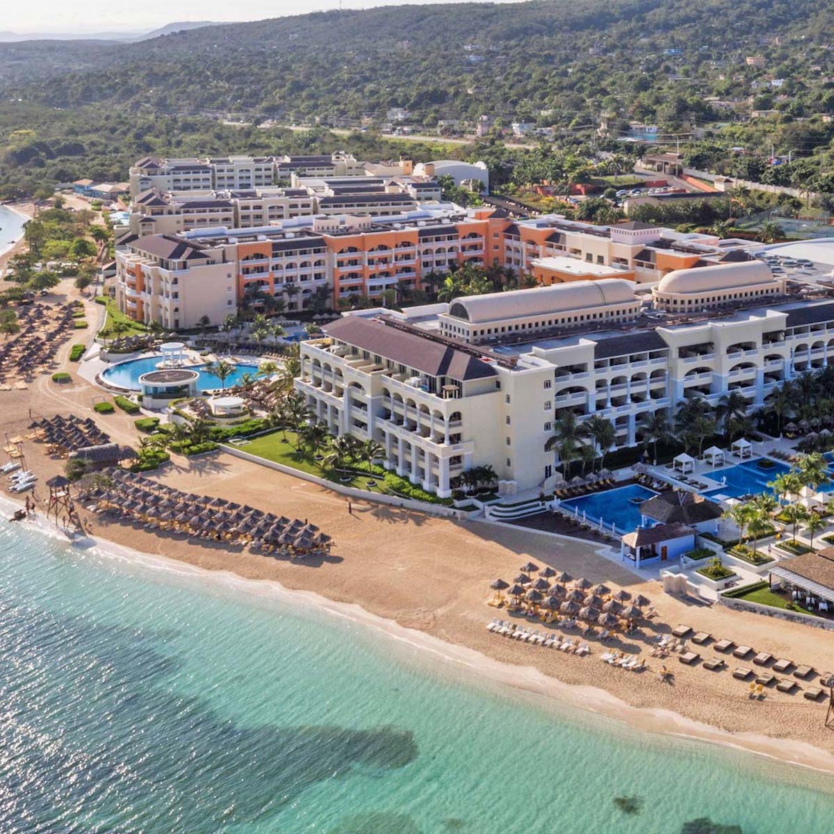 Aerial view of Iberostar Selection Rose Hall Suites, a large beachfront resort featuring multiple buildings, pools, palm trees, and loungers on the sandy beach by clear turquoise water.